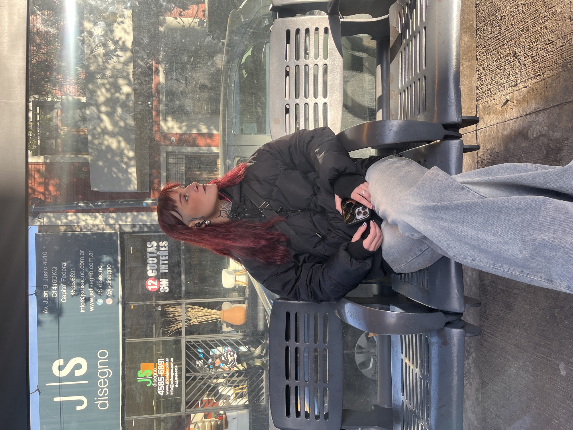 The image shows a young woman with red hair sitting on a gray metal bench at a bus stop or similar waiting area. She is wearing a black puffer jacket and gray jeans, and holding a cell phone in her hands. The background features a glass facade with advertisements for "JS Diseño," a design store. The text on the storefront provides the location as Av. Juan B. Justo 4910, C1416DKQ, Capital Federal, Buenos Aires, Argentina. The glass reflects the surroundings, including trees and buildings, suggesting an urban environment. The bench is attached to other seats of the same design. The lighting indicates it is daytime with possibly harsh sunlight.