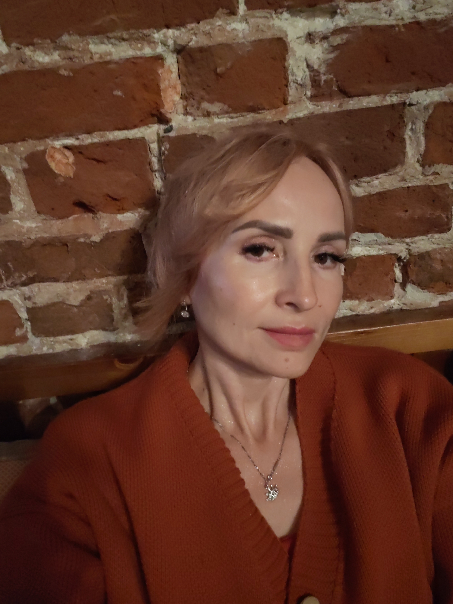 The media file is a medium close-up selfie of a woman with blonde hair, wearing an orange sweater and a silver necklace with a pendant. She is looking at the camera with a neutral expression. The background is a brick wall. The lighting suggests an indoor setting, possibly in the evening. The woman appears to be in Nizhny Novgorod, Russia. There are no visible text elements in the image.