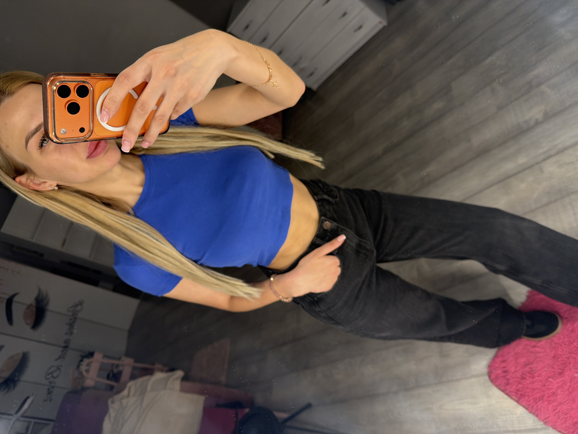 A blonde woman stands in front of a mirror, taking a selfie with her orange-cased phone. She is wearing a blue crop top and black jeans. Her left hand is in her pocket, and her right hand holds the phone. A pink rug is visible on the floor. In the reflection of the mirror, a shelf unit and a pink trolley can be seen. The environment suggests a room in a commercial establishment, possibly a salon, given the reflection of eyelash graphics on a mirrored surface in the background. The floor appears to be laminate wood. The overall lighting is moderate.