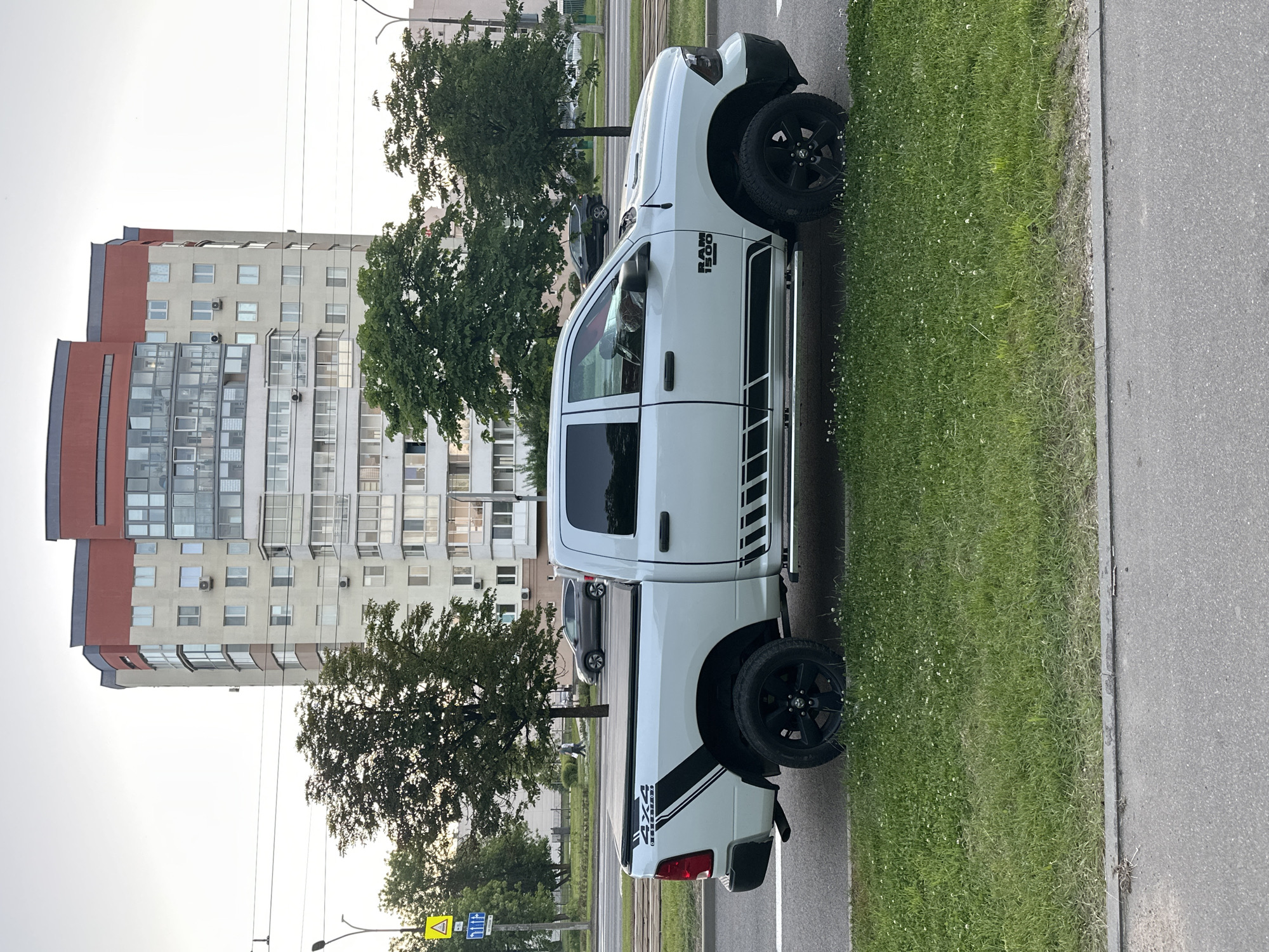A white Dodge Ram 1500 pickup truck is parked on the side of an urban road. The truck features black wheels, black side steps, and distinctive black striped decals on its front fender, displaying "RAM 1500," and on its bed, showing "4X4." It is positioned alongside a strip of green grass, which borders the asphalt road.
In the background, a tall, multi-story residential building with a contemporary design, characterized by grey, red, and extensive glass facades, rises under a bright, possibly overcast sky. Several trees with dense green foliage are visible between the truck and the building. A black sedan is partially seen further down the road, and a lone person can be observed walking in the distance behind the truck.
Overhead power lines and street light poles are present. Roadside signage includes a yellow triangular warning sign with a black exclamation mark and a blue rectangular sign featuring white symbols and Cyrillic text. The lighting suggests it is late afternoon or early evening. The scene is consistent with a residential area in Minsk, Belarus.