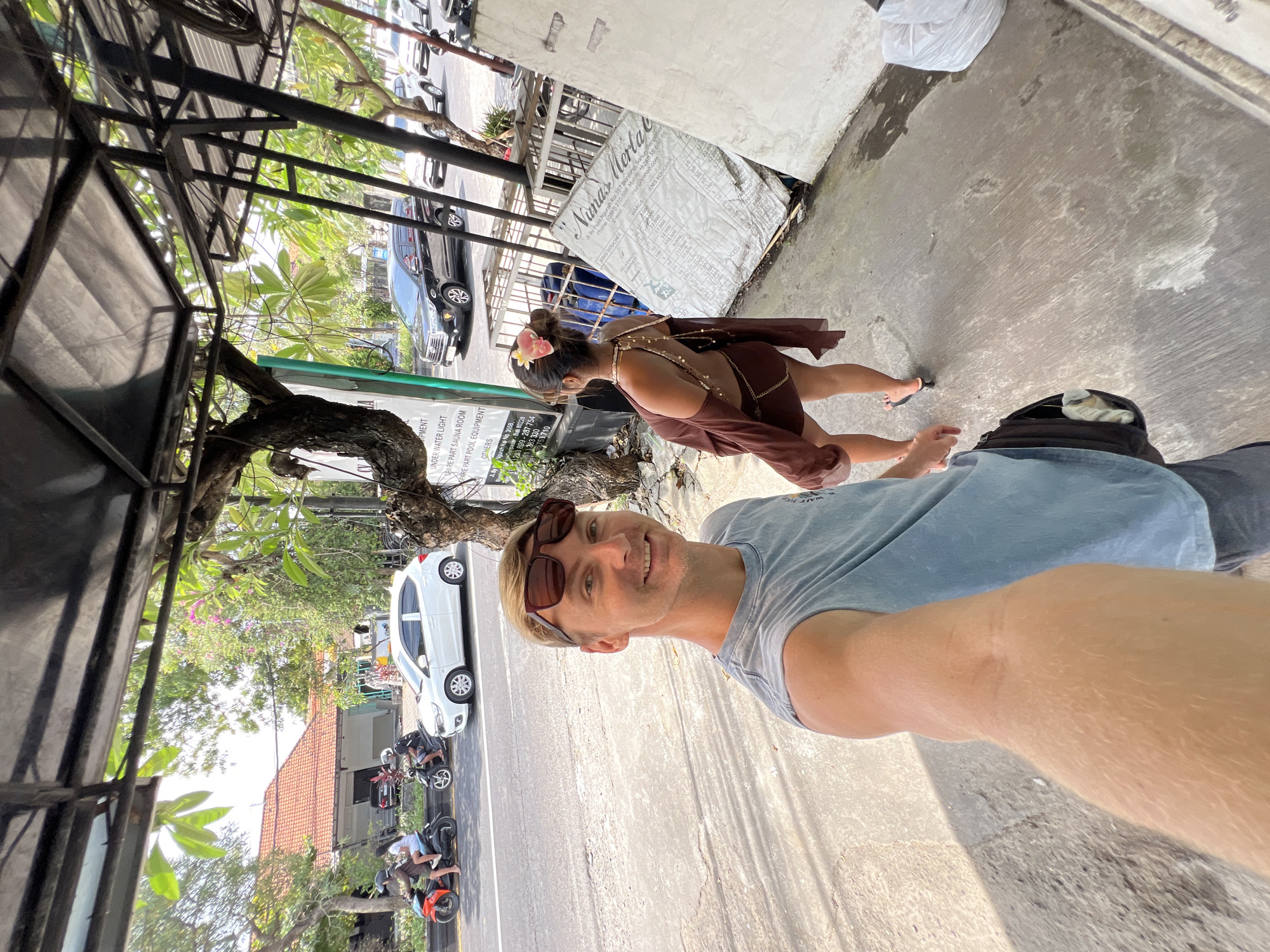 A man is taking a selfie in Denpasar, Indonesia. He is wearing a blue t-shirt and has sunglasses on his head. A woman with a flower in her hair is standing behind him. There is a street with cars and motorcycles in the background. There is a sign for "Nunas Merta" in the background.