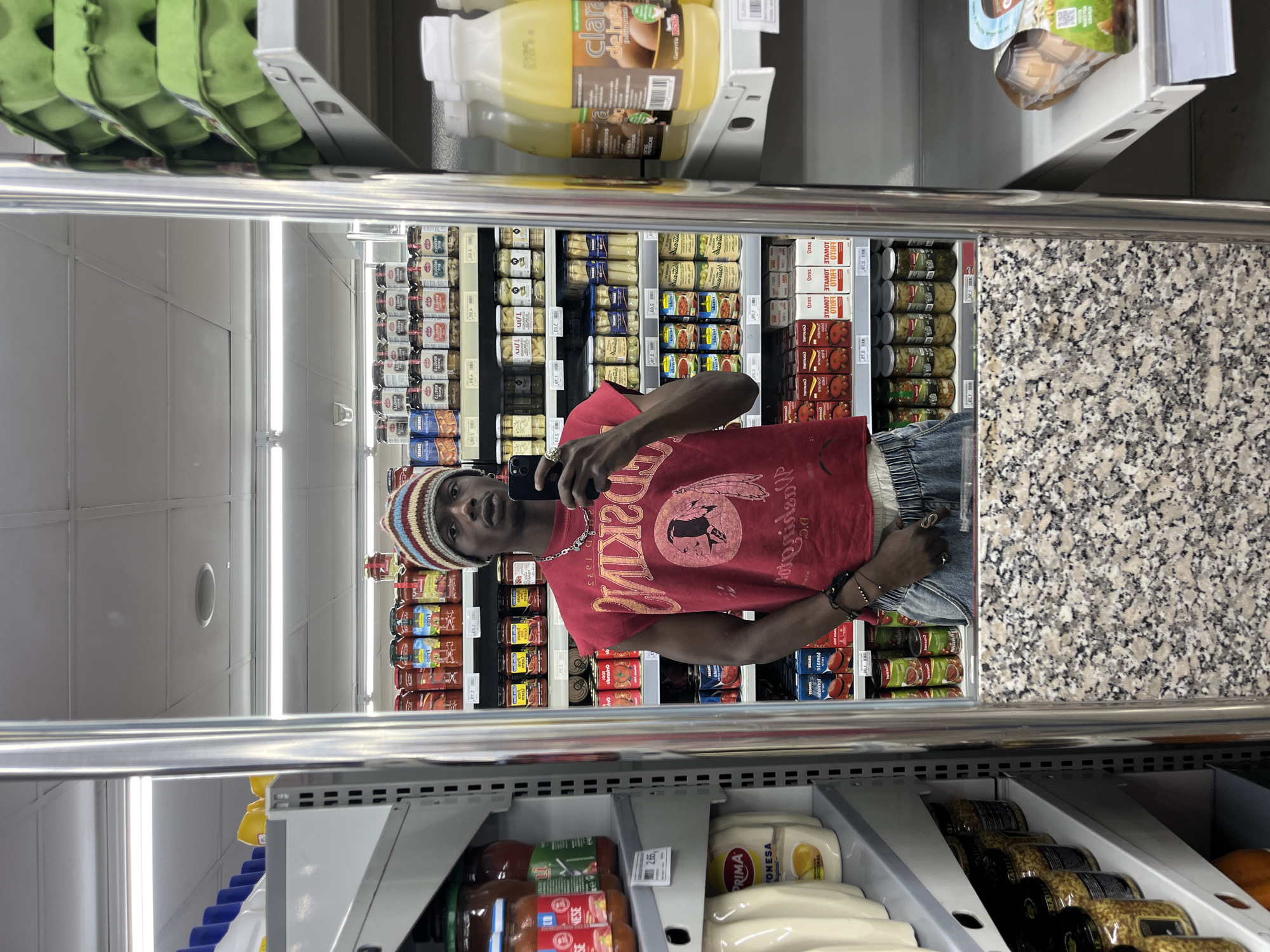 This portrait-oriented image, originally rotated 90 degrees clockwise, captures a young Black man taking a mirror selfie inside a supermarket in Barcelona, Spain. The man is reflected in a large, vertical mirror, which appears to be the door of a refrigerated display unit. He wears a red t-shirt with a graphic featuring a Native American head and the text "REDSKIN D.C.", blue jeans, a silver chain necklace, a black bracelet, and a multicolored striped knit beanie (red, orange, brown, beige, blue). He holds a black smartphone up, seemingly focused on the camera to capture his reflection.
The background, visible through the reflection, displays numerous supermarket shelves stocked with various goods. Canned tomatoes, with visible "TOMAT" and "ETA" labels, are prominent, alongside jars of olives and other preserves. Price labels such as "2,65", "0,95", and "1,20" are discernible on the shelves. Bright overhead fluorescent lighting illuminates the scene.
Outside the reflection, above the mirror, green egg cartons and bottles labeled "Clara de huevo pasteurizada" (pasteurized egg white) and "PROTICAS" are stacked. Below the mirror, shelves hold jars of tomato sauce and "PRIMA MONESA" (Prima Mayonnaise), indicating local Spanish brands. A speckled grey and white granite-like surface is visible on the right side within the reflection. The overall setting is a typical, well-lit grocery store.