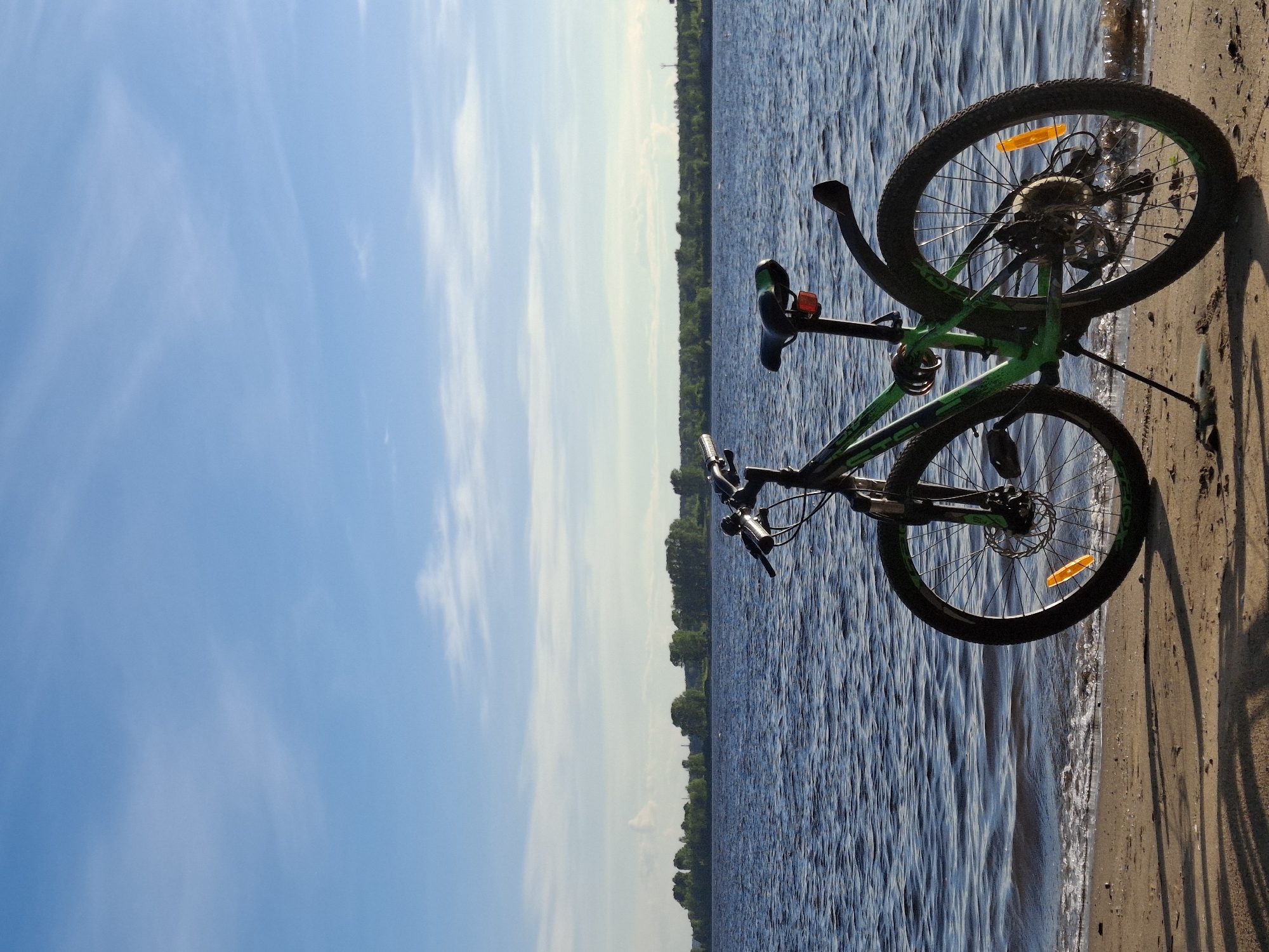 A green and black mountain bike is shown resting on a sandy shore next to a body of water. The bike is leaning against something, possibly another object or the ground, with its wheels facing upwards and downwards respectively. The front wheel is closer to the viewer and the handlebars are visible. The rear wheel, with a prominent orange reflector, is further away. The bike's frame features the word "GT" printed on it.
The scene appears to be a natural setting, likely a beach or riverbank. The water has ripples, suggesting a gentle breeze. In the background, a lush green tree line runs along the horizon, with a clear blue sky above, dotted with wispy white clouds. The overall impression is one of a serene and peaceful outdoor environment, possibly during a sunny day. The lighting suggests it might be late afternoon or early evening, given the long shadows cast on the sand. The location is identified as Chaikovsky, Russia. There are no people visible in the image, and the focus is solely on the bicycle and its surroundings.
