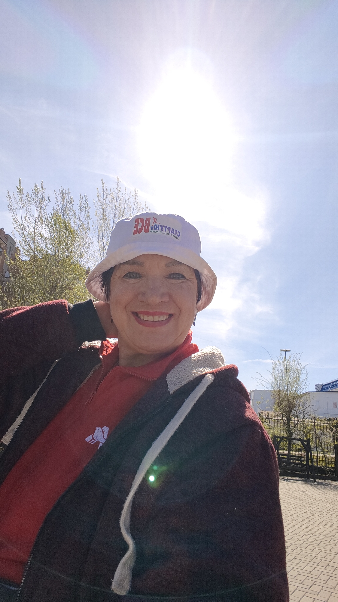 A woman is taking a selfie on a sunny day. She is wearing a white bucket hat with the Cyrillic inscription "START" and "BCE", a red zip-up hoodie with a small white logo, and a dark maroon jacket with white trim. She is smiling, and her right hand is touching the back of her neck. The background shows a blue sky with the sun shining brightly, some trees with budding leaves, a fence, a light pole, a building with signage in the distance, and a paved sidewalk. Based on the writing on the hat, this image was likely taken in the city of Krasnoyarsk, Russia.