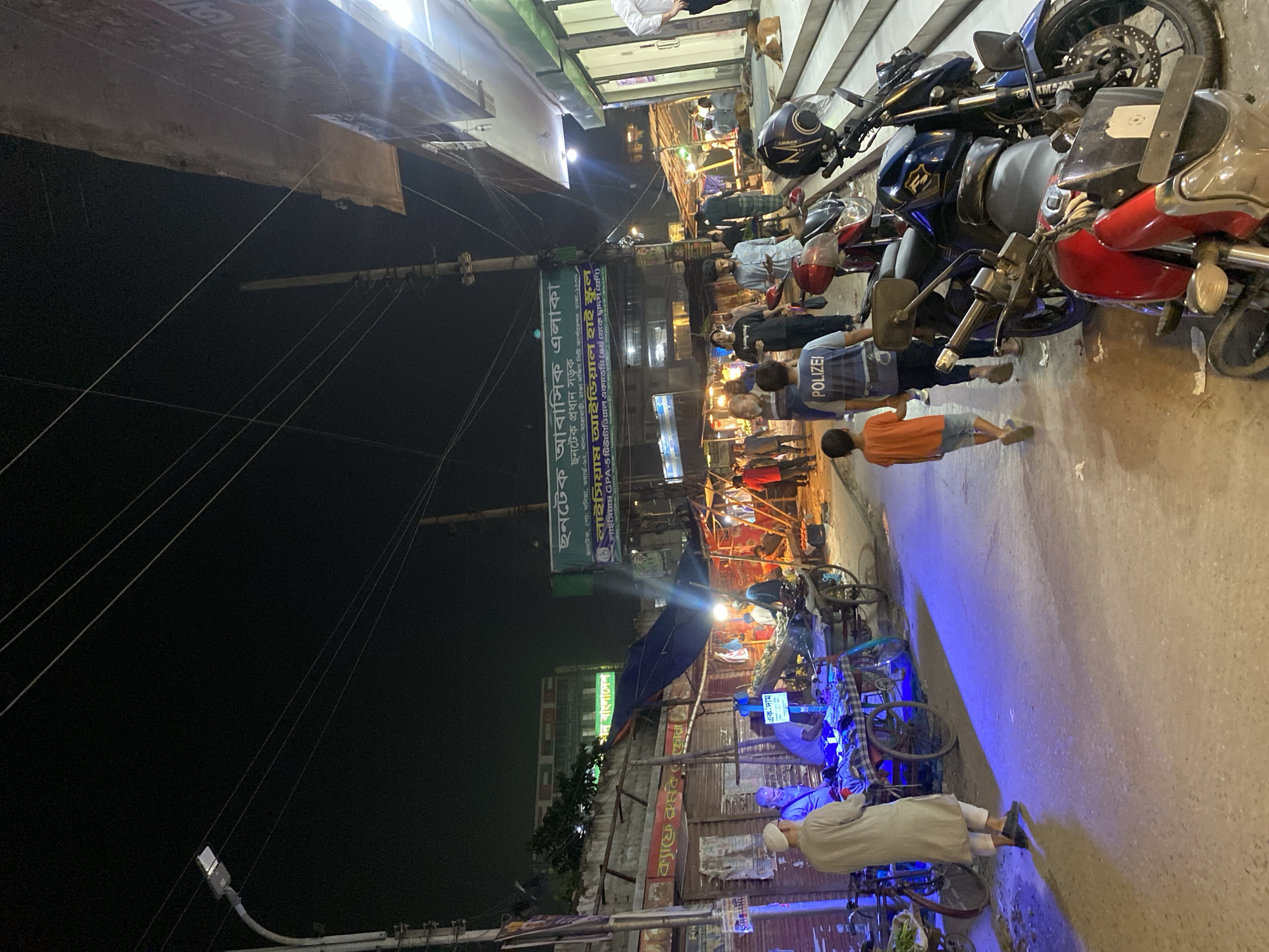 This image depicts a street scene at night in Dhaka, Bangladesh. The foreground shows a paved street lined with parked motorcycles and bicycle rickshaws. Pedestrians are walking along the street, including a person wearing a "POLIZEI" shirt, an orange-shirted child, and an older man in traditional attire.
Above the street level, there are businesses and signs. One sign is visible, advertising residential areas and an ideal high school. Power lines crisscross the sky. The image appears to be taken from a slightly elevated position, capturing the bustling activity of the urban environment.