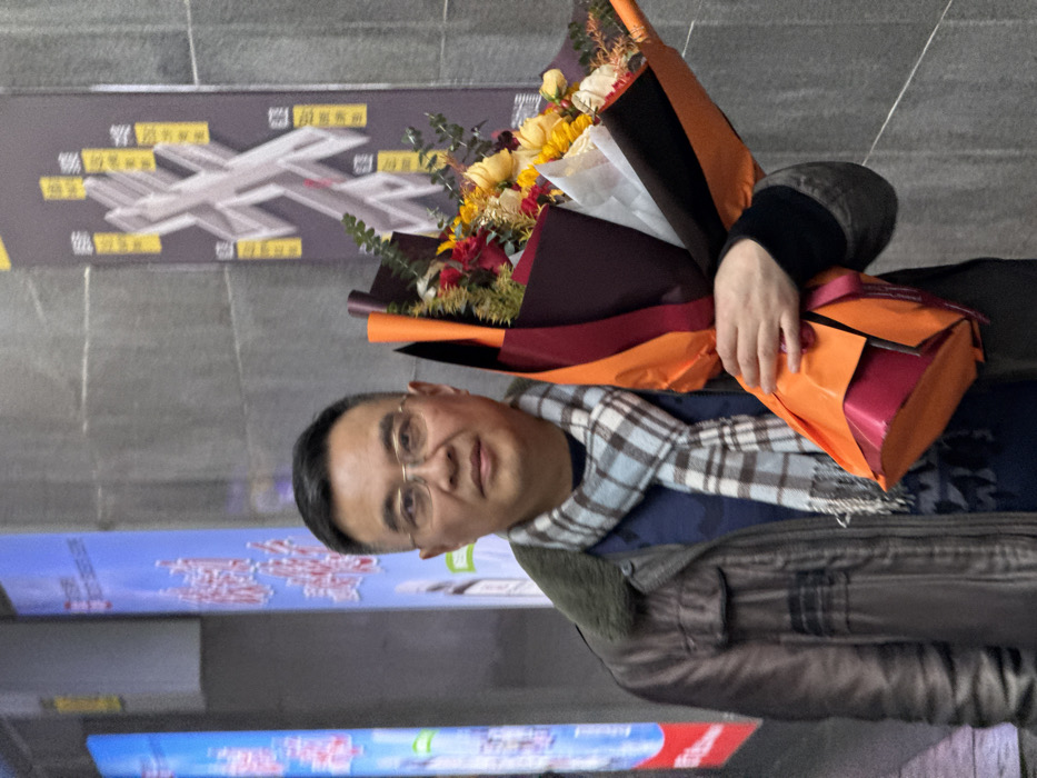 An East Asian male, appearing middle-aged, is depicted standing in what appears to be an urban indoor environment, likely within Yuzhong District, China. He is wearing eyeglasses, a dark brown jacket with a dark fur collar, a dark blue shirt or sweater, and a light blue and white plaid scarf with fringed ends. He holds a large floral bouquet in his left arm, wrapped in layers of orange, dark burgundy, black, and white paper, containing yellow and red roses and green foliage. A small magenta gift box, partially wrapped in orange paper, is held beneath the bouquet with his left hand. The man looks directly at the camera with a neutral expression. The background consists of dark grey tiled walls. To the left, a vertical illuminated display board shows white Chinese characters on a blue background. To the right, a vertical poster features a stylized metallic 'X' design surrounded by yellow Chinese text on a grey background. Artificial lighting illuminates the scene.
