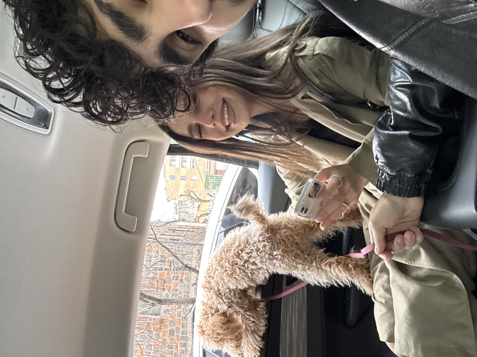 The image captures a selfie within a vehicle, likely a car, taken by a male and female. The male figure is positioned at the top left, with curly dark hair and dark eyes visible. The female is adjacent to him, smiling and looking at the camera while holding a mobile phone.
Between them is a light-brown curly-haired dog, possibly a poodle mix, with a pink leash, positioned towards the car window and a view of the exterior. The woman seems to be holding the leash. The car window looks out to a scene with building exteriors and trees, suggesting an urban setting.
Based on the reference to Yerevan, Armenia, the urban environment may be located within this city.