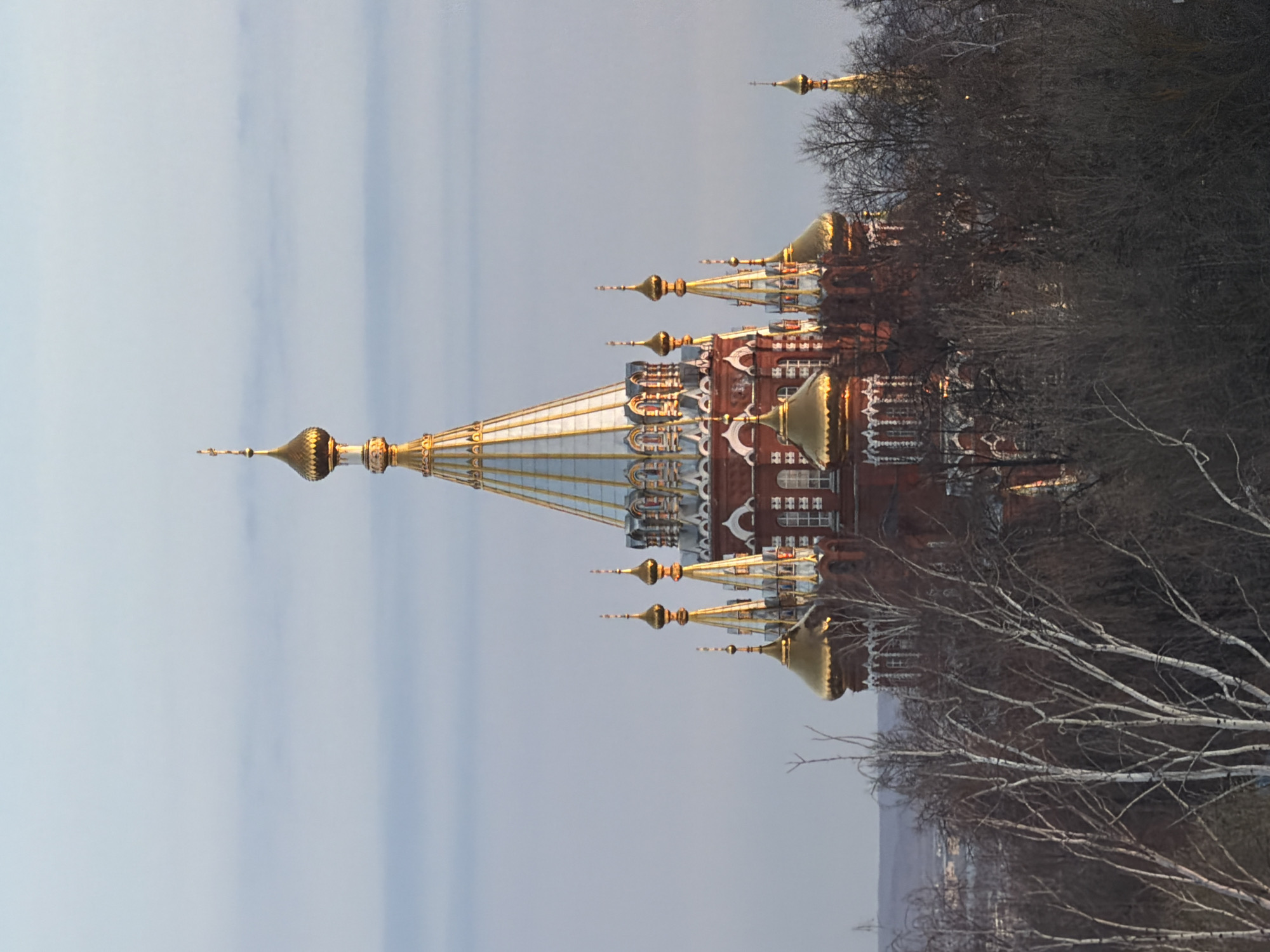 The image shows the Orthodox St. Michael's Cathedral in Izhevsk, Russia. The cathedral, with its red brick walls, white trim, and gold domes, is partially obscured by bare trees on the right. The sky is overcast.