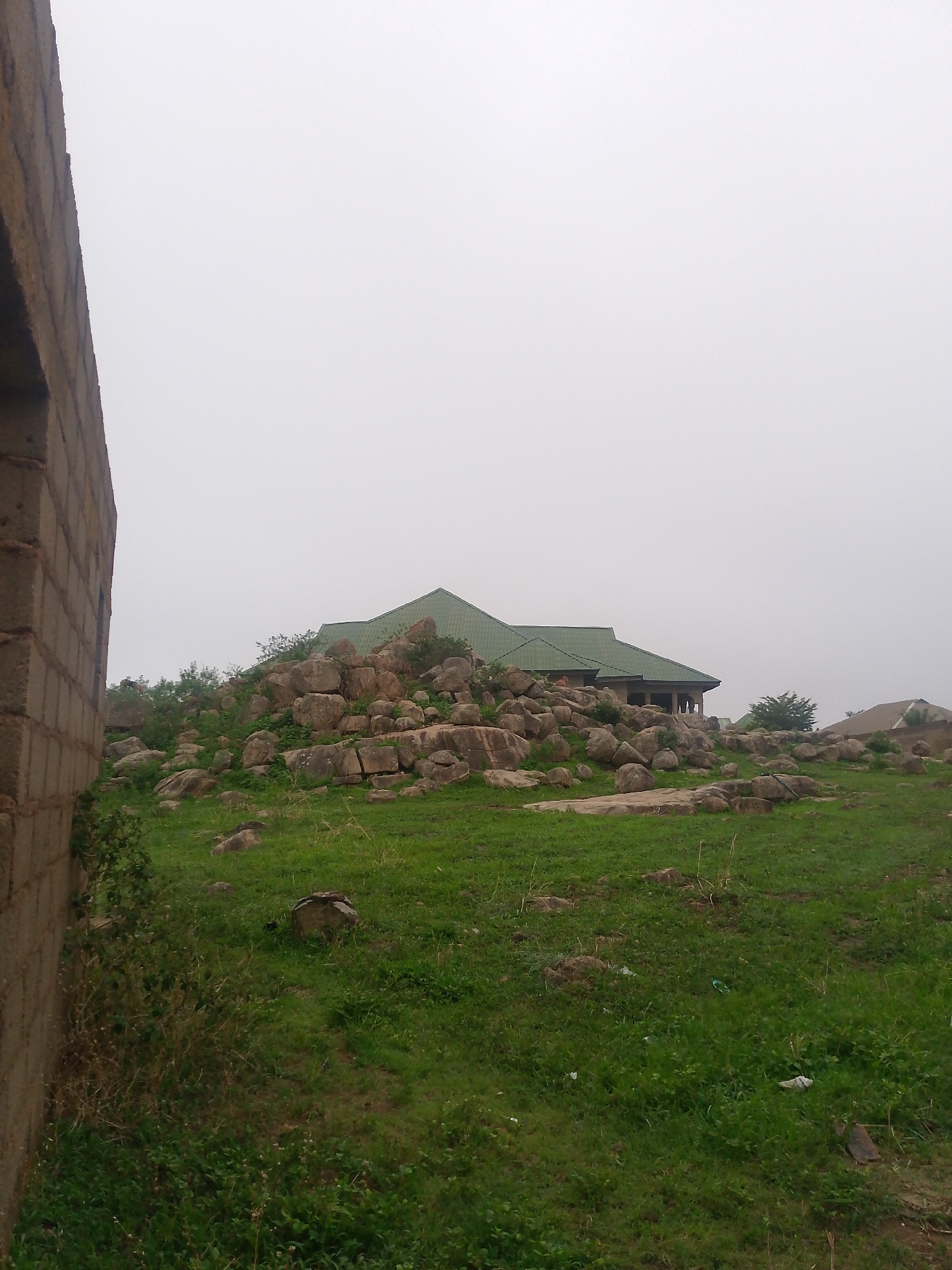The image depicts a green, grassy landscape in Barikin Gidar Kanat, Nigeria. A house with a green roof is situated on top of a rocky hill, with large boulders scattered across the slope. The sky is overcast and pale. The corner of a brick structure is visible on the left side of the frame.