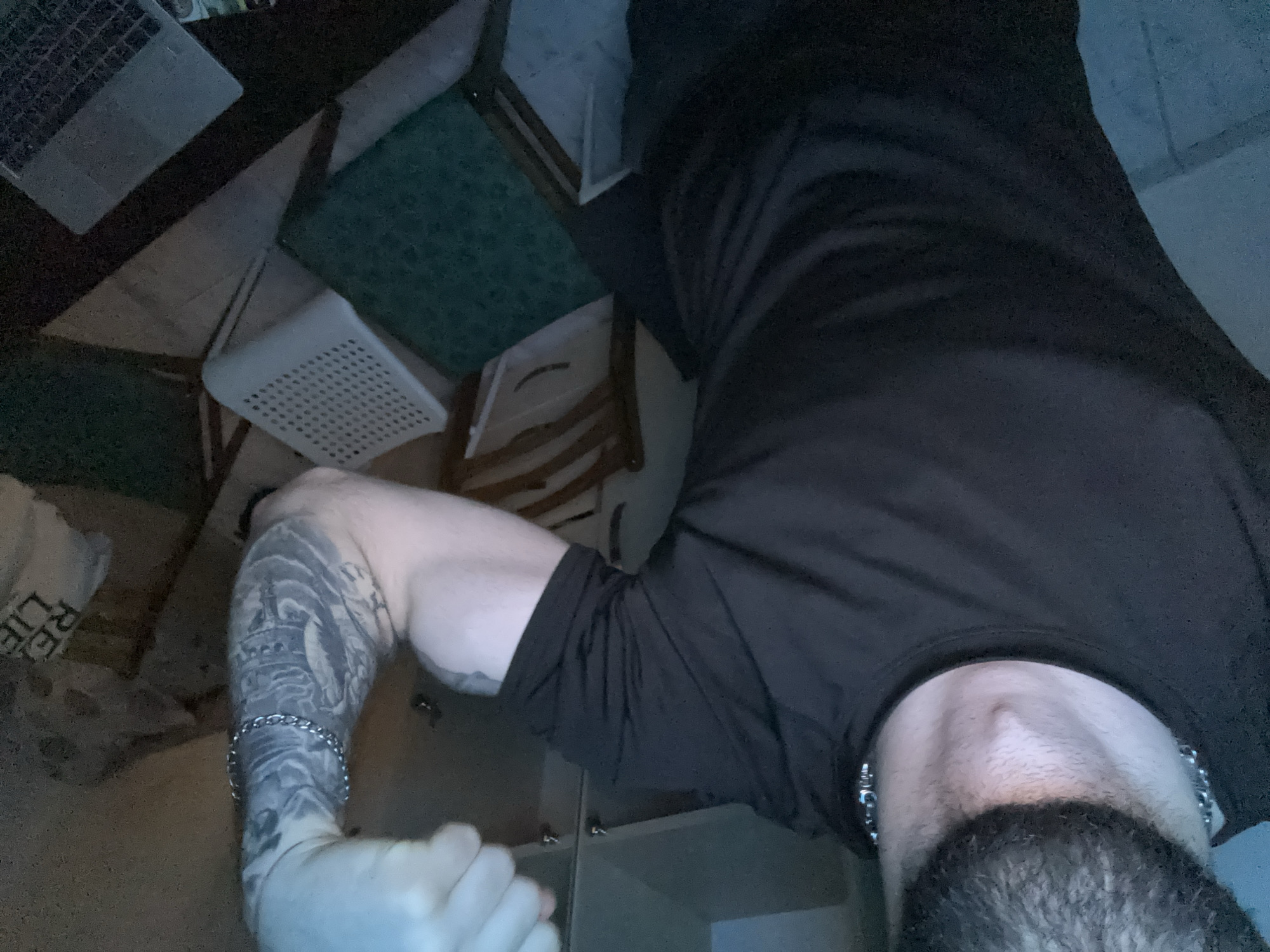 The image shows a close-up view of a person's upper body, with a focus on their bicep. The person is wearing a dark t-shirt and a silver chain around their neck. Their left arm is bent, showcasing a bicep with extensive tattoos covering it. The tattoos are in grayscale and depict various images, including what appears to be a helmet and possibly figures. A silver bracelet is visible on the tattooed arm, near the wrist.
The background is dimly lit and appears to be an indoor setting. Several chairs are visible, some with dark frames and others with white plastic seats. A green cushioned seat is also present. In the far background, there seems to be some furniture or cabinetry. A laptop is partially visible in the lower right corner of the image. The lighting suggests the image was taken at night or in a poorly lit room. The scene does not provide any specific clues to pinpoint the exact location within Parvomay, Bulgaria.