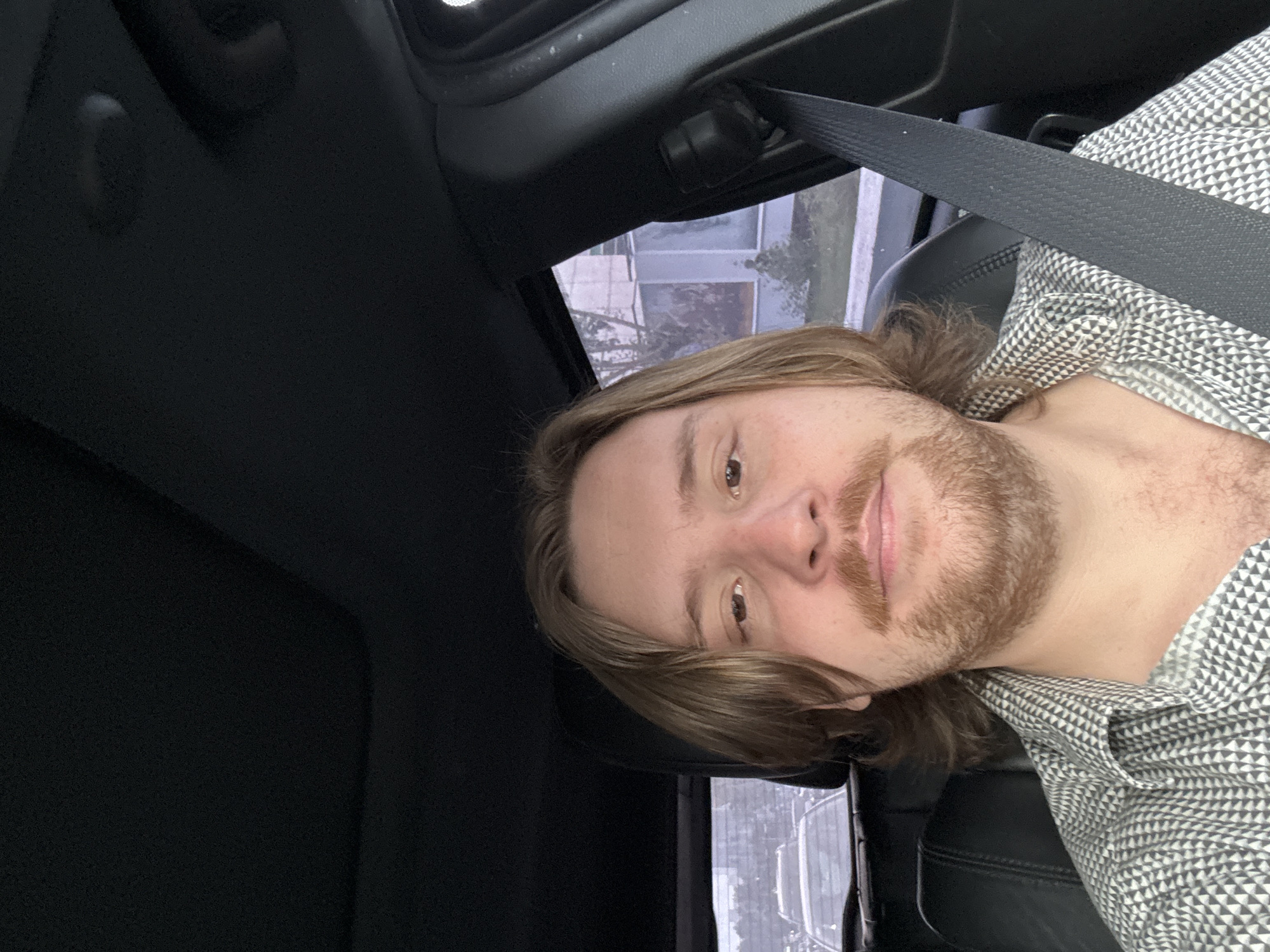 A man with light brown hair and a beard is sitting in the driver's seat of a vehicle, wearing a gray and white patterned shirt and a seatbelt. He is looking directly at the camera. The interior of the car is mostly black. Outside the car window, part of a building can be seen. The photo appears to be taken from inside the car. The location is reported to be Mexico City, Mexico.