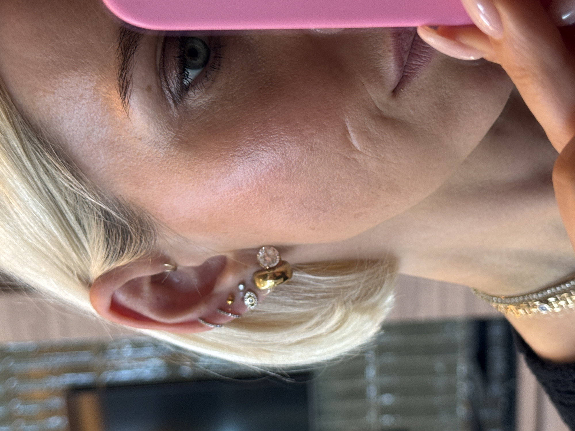 A close-up, sideways shot captures the right side of a person's head, revealing blonde hair, fair skin, and a visible eye. A pink object, likely a smartphone, is partially obscuring the top of the face, held by a hand with long, light pink/nude manicured nails. The person's left eye, featuring a dark iris and visible lashes, is partially visible beneath the pink object.
The ear is adorned with multiple piercings. In the lobe, there are several small studs, including a gold-toned one and one with a clear stone. Further up, a larger, irregular gold-toned piece is visible, along with another large, round clear-faceted stone earring. A small silver hoop is seen along the helix of the ear, and another silver-toned curved barbell or hoop is present further down the cartilage. Blonde hair is styled around the ear.
The skin on the face shows visible pores and fine lines, particularly around the eye and cheekbone. A faint line or scar is present on the cheek area. Below the jawline, a sparkling, gold-toned accessory, possibly a necklace or bracelet with small stones, is visible. The background is blurred, suggesting an indoor setting. No specific activities beyond holding the object close to the face and no discernible text are visible. The overall impression is a detailed, candid portrait focusing on facial features and accessories.