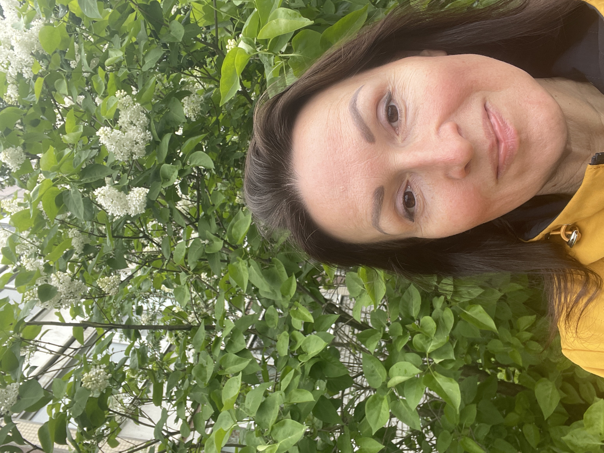 A woman with brown hair, wearing a yellow jacket, is standing in front of a lilac bush with white flowers. The picture was taken outdoors, and a building is visible in the background. The location is Saint Petersburg, Russia.