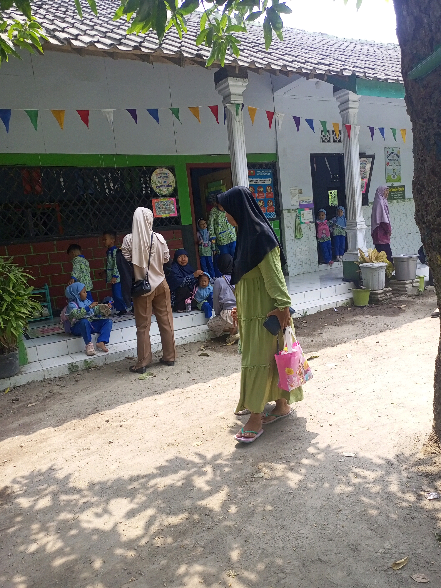 A street scene in Gurah, Indonesia, shows a group of children and adults outside a building with a tiled roof and white walls adorned with colorful triangular flags. The building appears to be a school or community center, with several posters displayed on the exterior. In the foreground, a woman in a long green dress and black hijab walks away from the camera, carrying a pink tote bag. Several children in school uniforms are scattered around the area, some sitting on steps and others standing. One adult, also wearing a hijab, is seated on the steps. The ground is a dirt path with shadows cast by overhead foliage.
