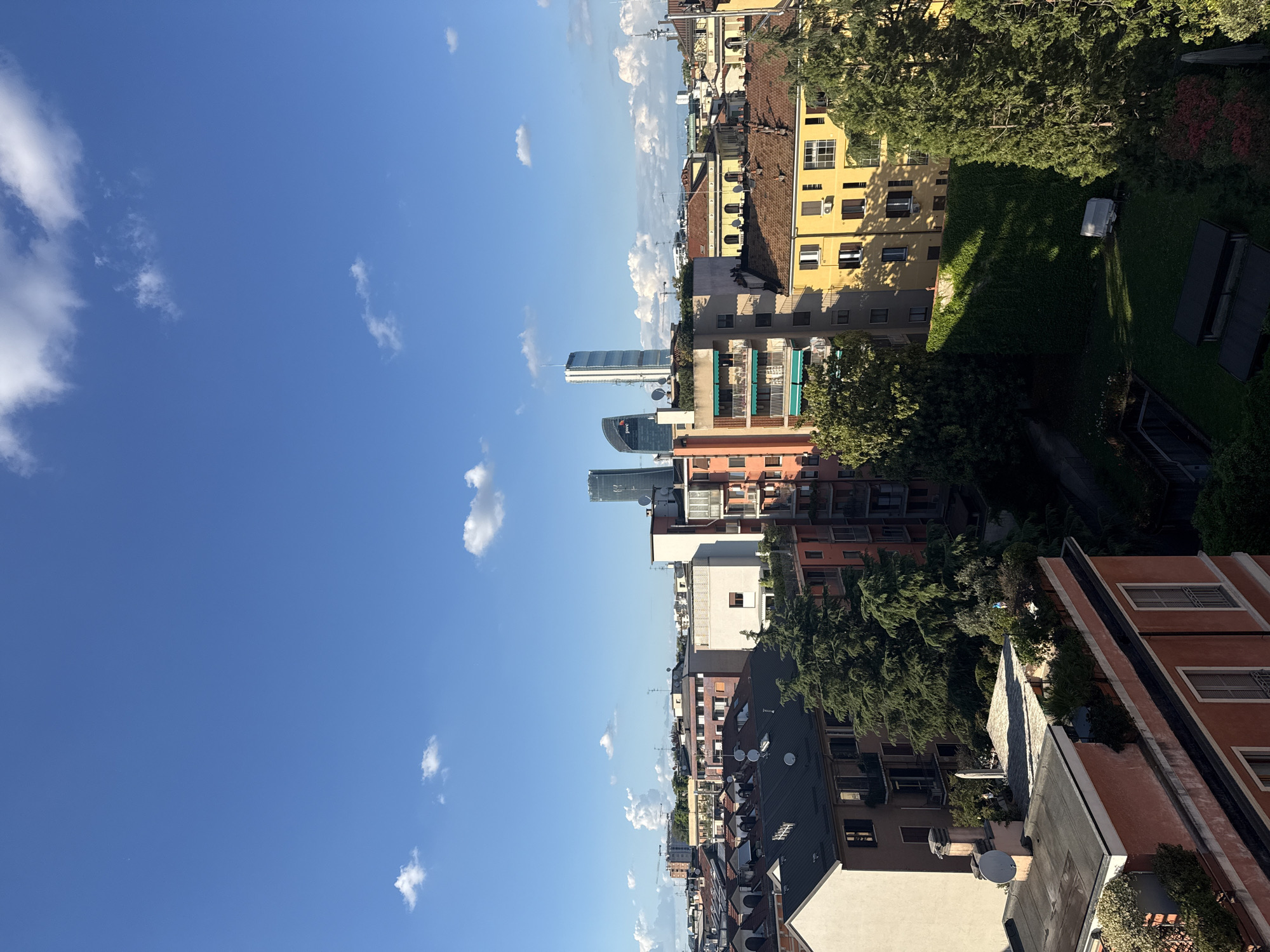 The image shows an elevated view of Milan, Italy, under a clear, blue sky with scattered clouds. The architecture consists primarily of mid-rise buildings of varying colors: yellow, red, white, and brown, with traditional rooftop structures. Modern skyscrapers are visible in the distance. Trees are interspersed among the buildings and gardens with ivy are present. There are no people visible in the image.