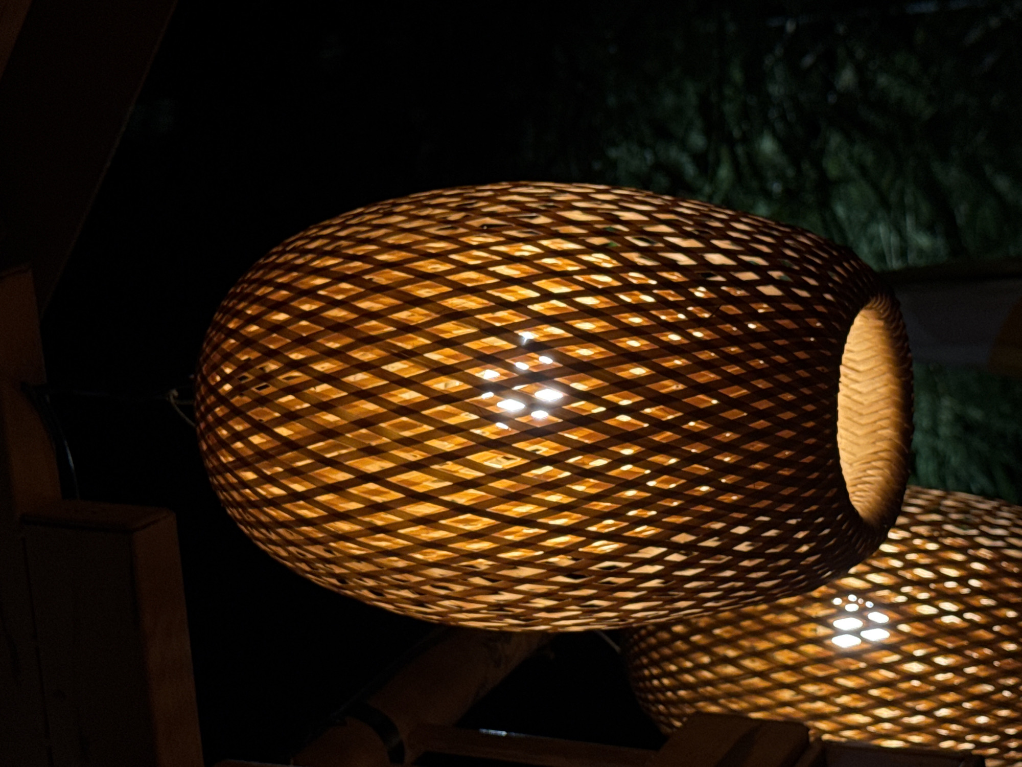 The image shows two woven bamboo lamps hanging in darkness. The lamp in the foreground is larger and in focus, revealing a diamond-patterned weave that allows light to escape from within. The light source inside the foreground lamp is visible as a bright, diffuse glow. Behind it, a second, smaller lamp is also visible, similarly constructed and illuminated, though it is out of focus. The lamps are suspended from a dark wooden structure. The background is dark, with hints of blurred green foliage suggesting an outdoor setting at night, potentially in a garden or courtyard. The overall ambiance is warm and inviting, with the light from the lamps creating a cozy atmosphere. There are no people present in the image. The location is not specified in the image or accompanying information.