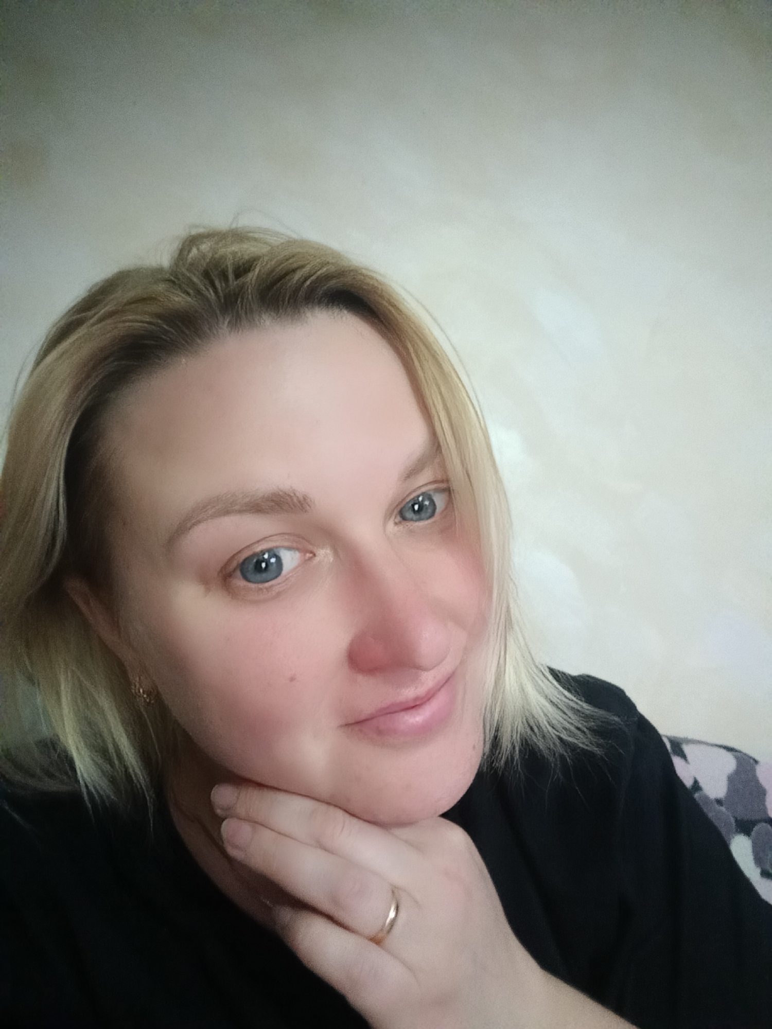 A blonde woman, likely located in Chaikovsky, Russia, is depicted in a head-and-shoulders portrait, presumably a selfie, taken indoors during daytime. She has fair skin, striking blue eyes, and natural-looking eyebrows. Her short to medium-length blonde hair is casually styled, framing her face. She is wearing a simple black top or t-shirt.
The woman is gently resting her right hand under her chin, with a plain gold band ring visible on her right ring finger. She looks directly at the camera with a calm, pleasant expression and a subtle, closed-mouth smile, conveying a serene demeanor.
The background is a light-colored, possibly off-white or beige wall that appears to have a slightly textured finish. To the far right, a small part of a patterned fabric, possibly from a piece of furniture or another garment, can be seen, featuring abstract shapes in shades of grey and possibly light pink. The lighting is soft and even, typical of an indoor setting. No text is visible in the image.