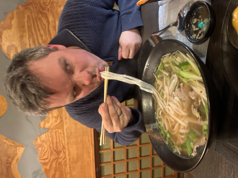 A person with graying hair and a blue sweater is eating a long white noodle using chopsticks. The person is looking at the camera with a slightly skewed expression. The noodle stretches from the chopsticks directly into the person's mouth. In front of the person is a large black bowl filled with broth, rice noodles, bean sprouts, and green onions, consistent with Pho soup. A small dish with a dark sauce is visible near the soup, and the background has light brown wood paneling.