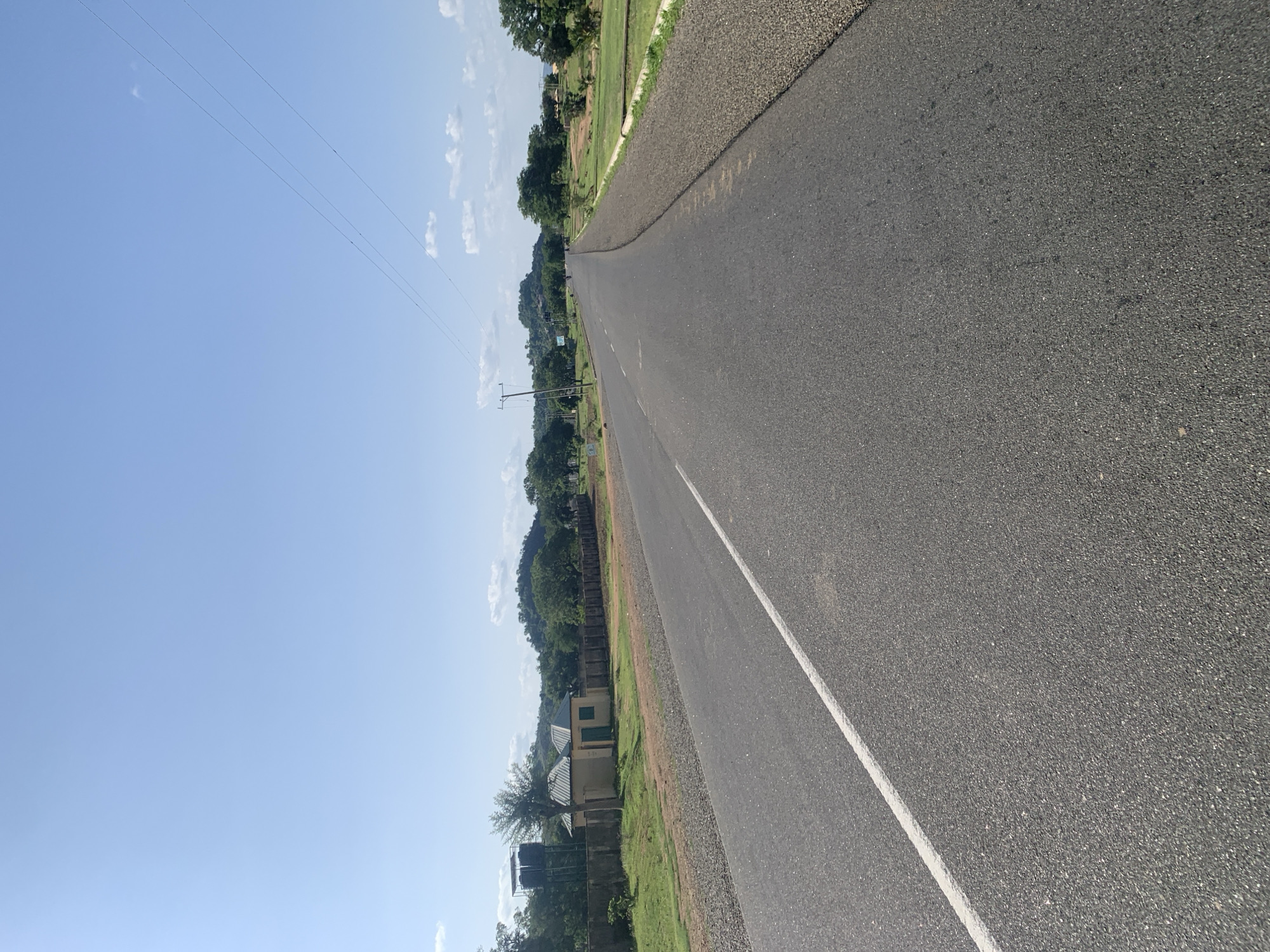An asphalt road stretches into the distance under a clear blue sky dotted with a few white clouds. A white line marks the left lane of the road. On the left side of the road, a concrete wall encloses a property with two small, light-colored buildings with green doors and windows, and two water tanks on a raised platform. Beyond the wall, green grass and trees are visible, with rolling hills covered in vegetation in the background. On the right side of the road, the shoulder is covered in gravel, and beyond that, there are grassy fields and scattered trees. Utility poles and power lines are present along the right side of the road. The scene appears to be in a rural or semi-rural area of Boi A, Nigeria, given the infrastructure and landscape. There are no people clearly visible in the image.