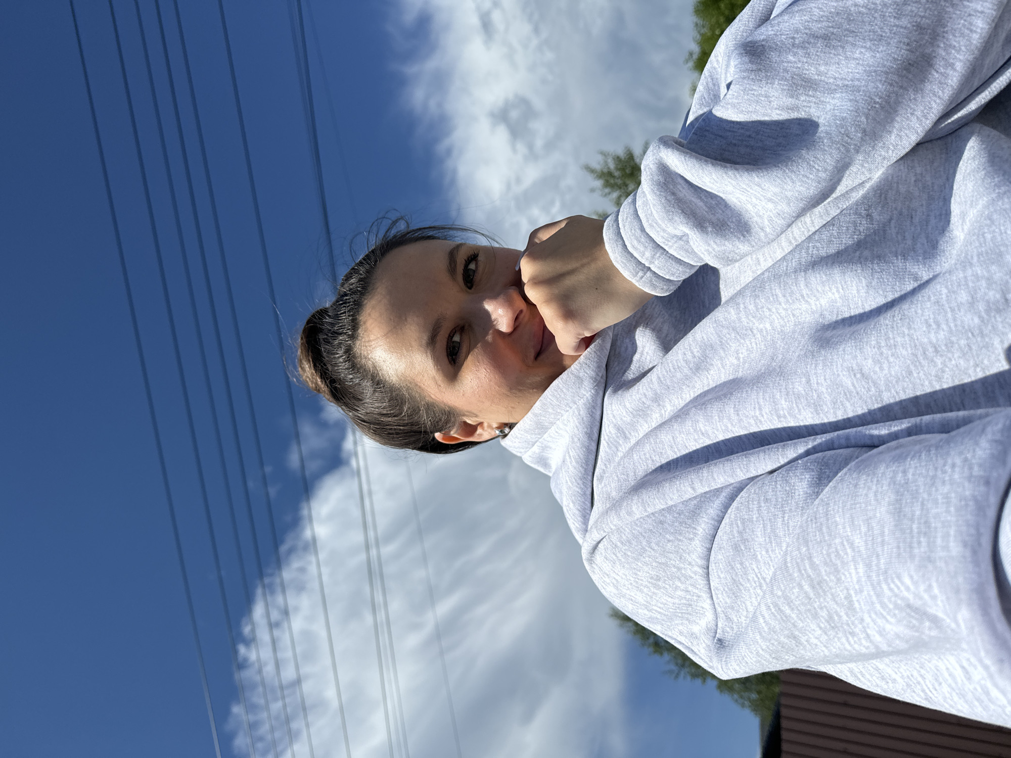 A woman with brown hair pulled into a bun is shown in a selfie against a partly cloudy sky. She is wearing a light grey hooded sweatshirt. Electrical power lines are visible in the upper left of the frame. The location is suggested to be in or near Vysokinichi, Russia, based on the request.