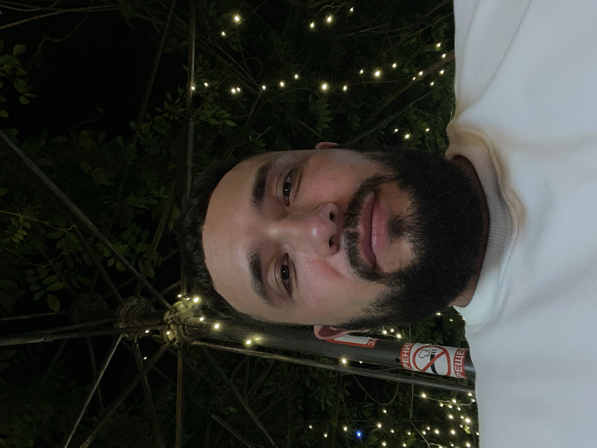 A man with a beard and a white sweatshirt is taking a selfie at night. He is standing near a pole with a "No Smoking" sign on it. The background is dark and there are string lights in the trees. The man is looking at the camera and smiling. The photo appears to have been taken in Makhachkala, Russia.