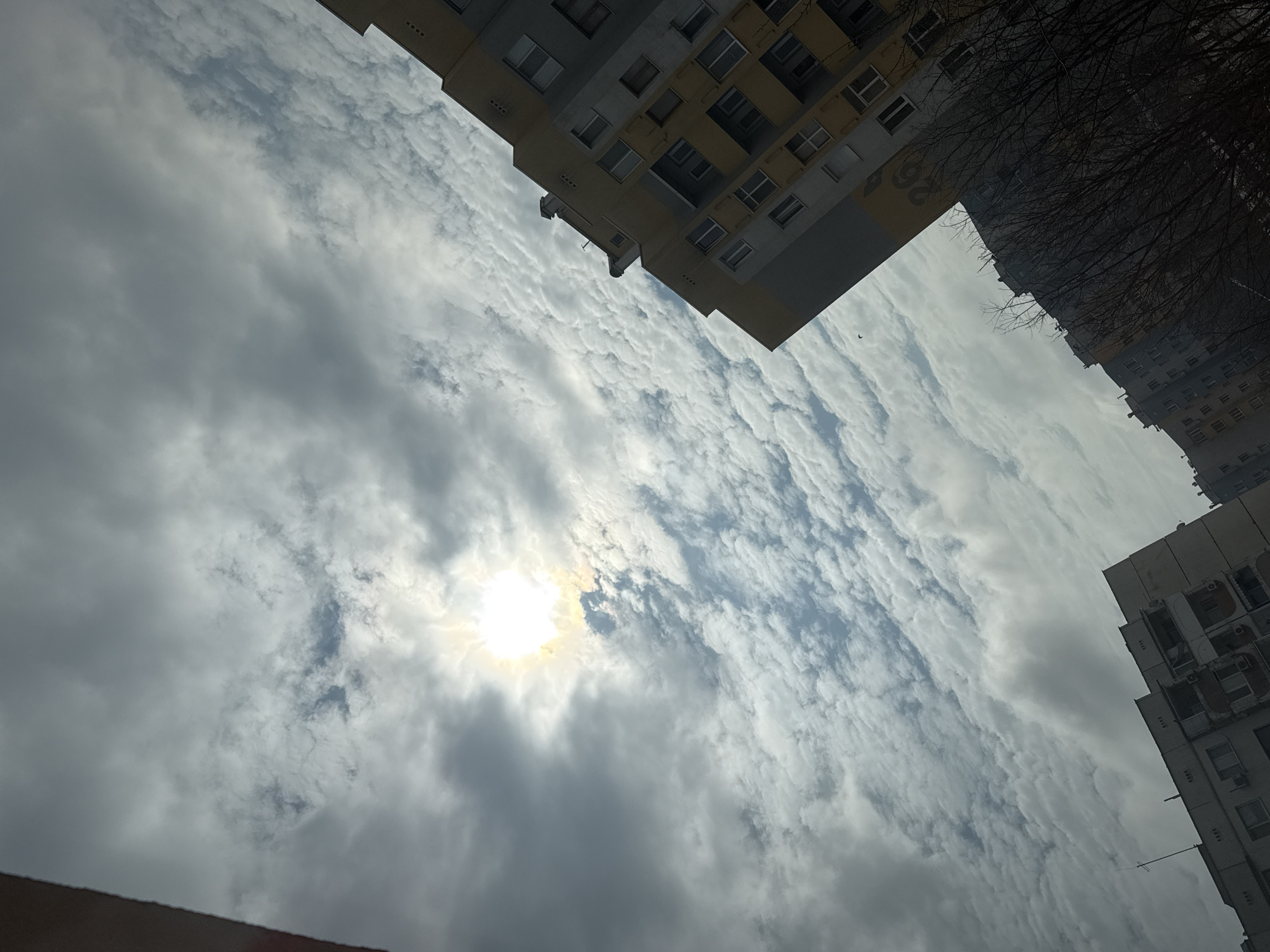 The image is a low-angle shot looking up towards the sky. The sky is predominantly filled with clouds, with the sun visible through a break in the cloud cover. Two buildings are partially visible on either side of the frame. The building on the left is a light yellow color. The building on the right is white. Bare tree branches are visible above the building on the right. The location is Stara Zagora, Bulgaria.