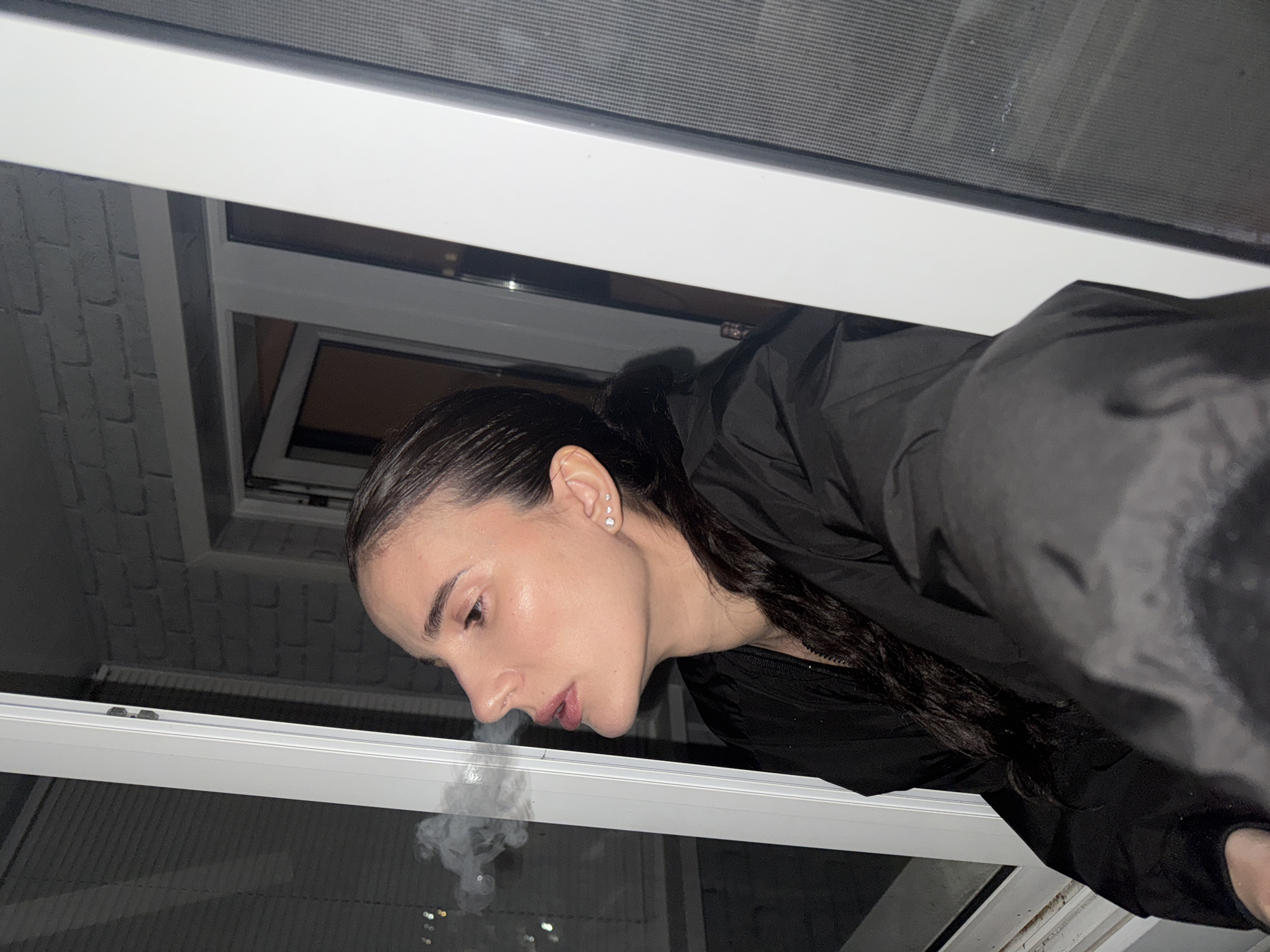 A low-angle shot shows a woman leaning out of a window. She has dark hair, slicked back, and is wearing a black jacket. Exhaled smoke is visible near her mouth. The window has a white frame and overlooks a cityscape at night, with faint lights visible in the distance. A brick wall is partially visible through an inner window. The location is stated as being in the city of Chaikovsky, Russia.