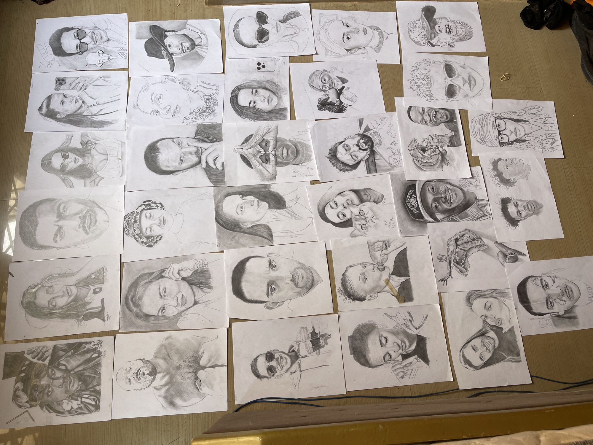 The image shows numerous portrait sketches on white paper scattered across a brown surface. The sketches appear to be done in pencil or charcoal, and feature a variety of subjects, including men and women, some wearing hats or glasses. Some sketches include additional elements like cats or other people. The environment appears to be an indoor space with possible floor coverings. There is no clear indication of Jos, Nigeria.