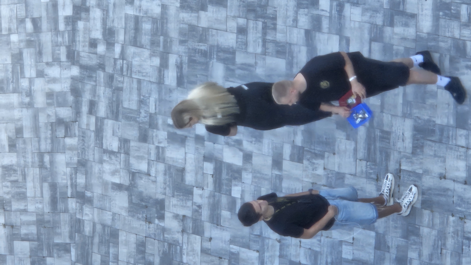 The image depicts three individuals standing on a patterned tiled surface. In the foreground, a young man with dark hair is seen from the waist down, wearing a black t-shirt and denim shorts, with his hands in his pockets. To his right, a woman with long blonde hair, wearing a black t-shirt and pants, is facing away from the viewer. To the right of the woman, another young man is visible from the waist down, wearing a black t-shirt and shorts. He is holding a blue container filled with small objects and a red container. The tiled surface appears to be an outdoor area, possibly a plaza or walkway. The overall lighting suggests daylight. Without further context or identifying markers, it is not possible to determine the specific location within the city of Prešov, Slovakia.