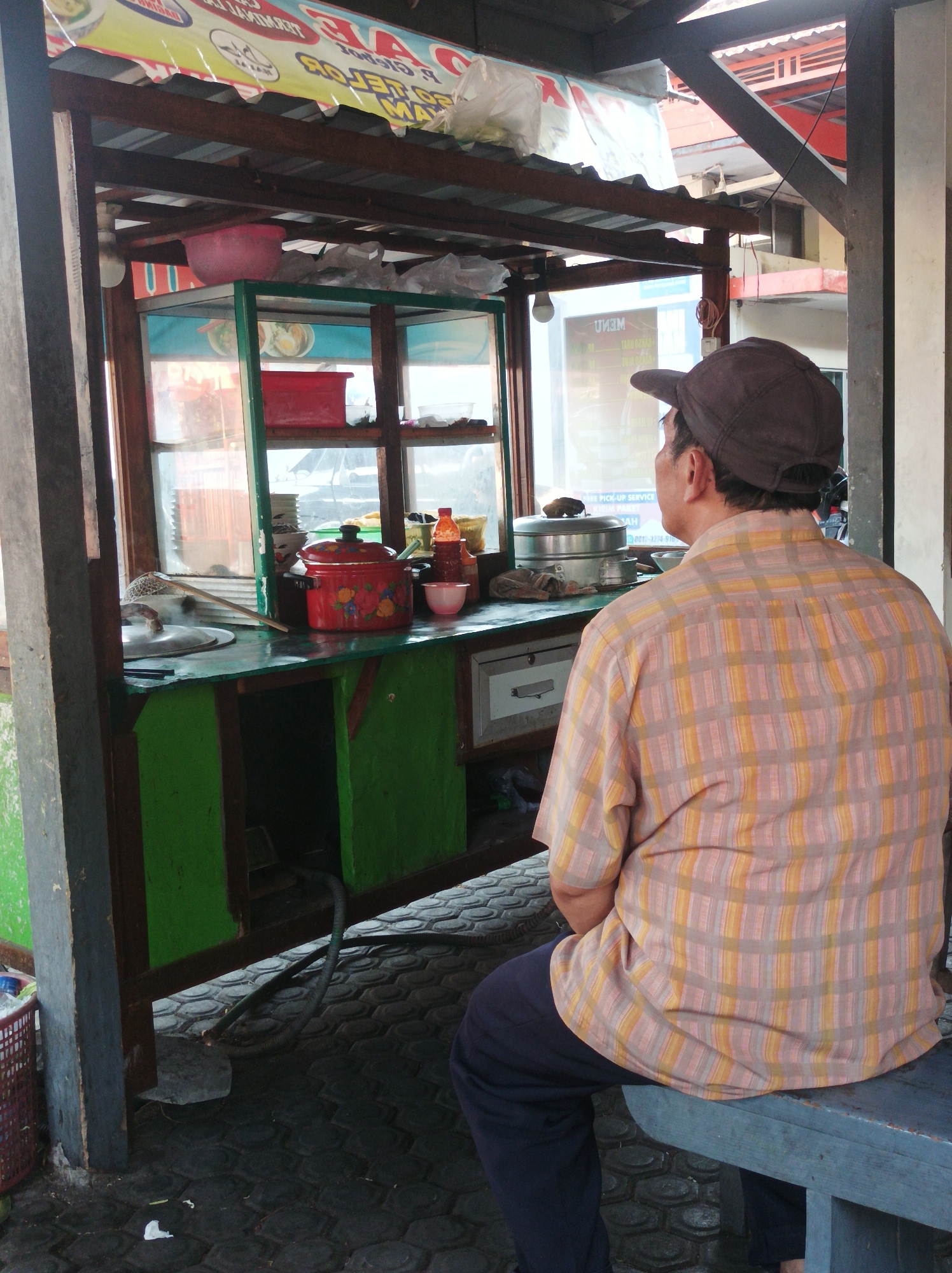 The image depicts an outdoor food stall in Nganjuk, Indonesia. A man wearing a plaid shirt and a cap sits on a blue bench, facing away from the camera. He appears to be observing the food stall, which features a display case with various items, including pots, containers, and what appear to be food ingredients. A red pot with a floral design is prominently displayed on the counter. The stall is constructed with wood and metal, and the ground is paved with interlocking hexagonal stones. A banner with text is visible above the stall, suggesting it is a business. The environment appears to be a street or alleyway, with buildings visible in the background.