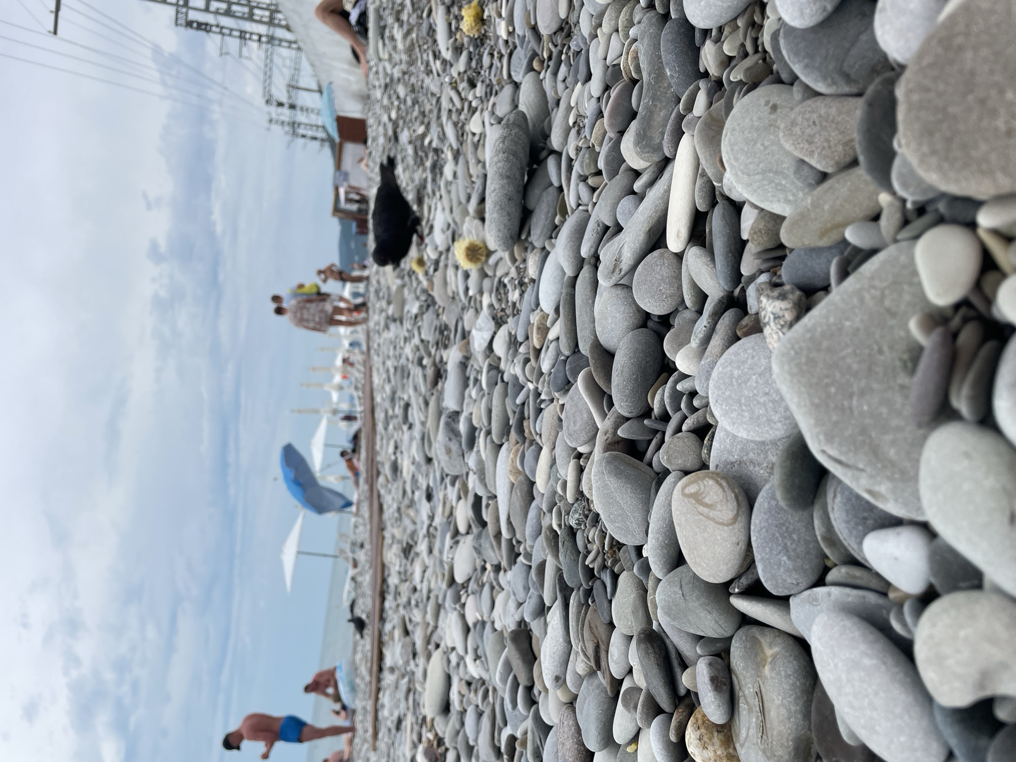 This is a low-angle, eye-level shot of a pebble beach on a cloudy day. In the foreground, the frame is filled with an abundance of smooth, gray and off-white pebbles of various sizes. A black bird is positioned in the mid-ground, pecking at something on the ground. Further in the background, several people are visible on the beach, some relaxing under umbrellas and others walking. There are also lounge chairs and a blue umbrella scattered across the beach. In the far background, the sea is visible, along with some distant structures and power lines. The overall impression is that of a busy, public beach on an overcast day.