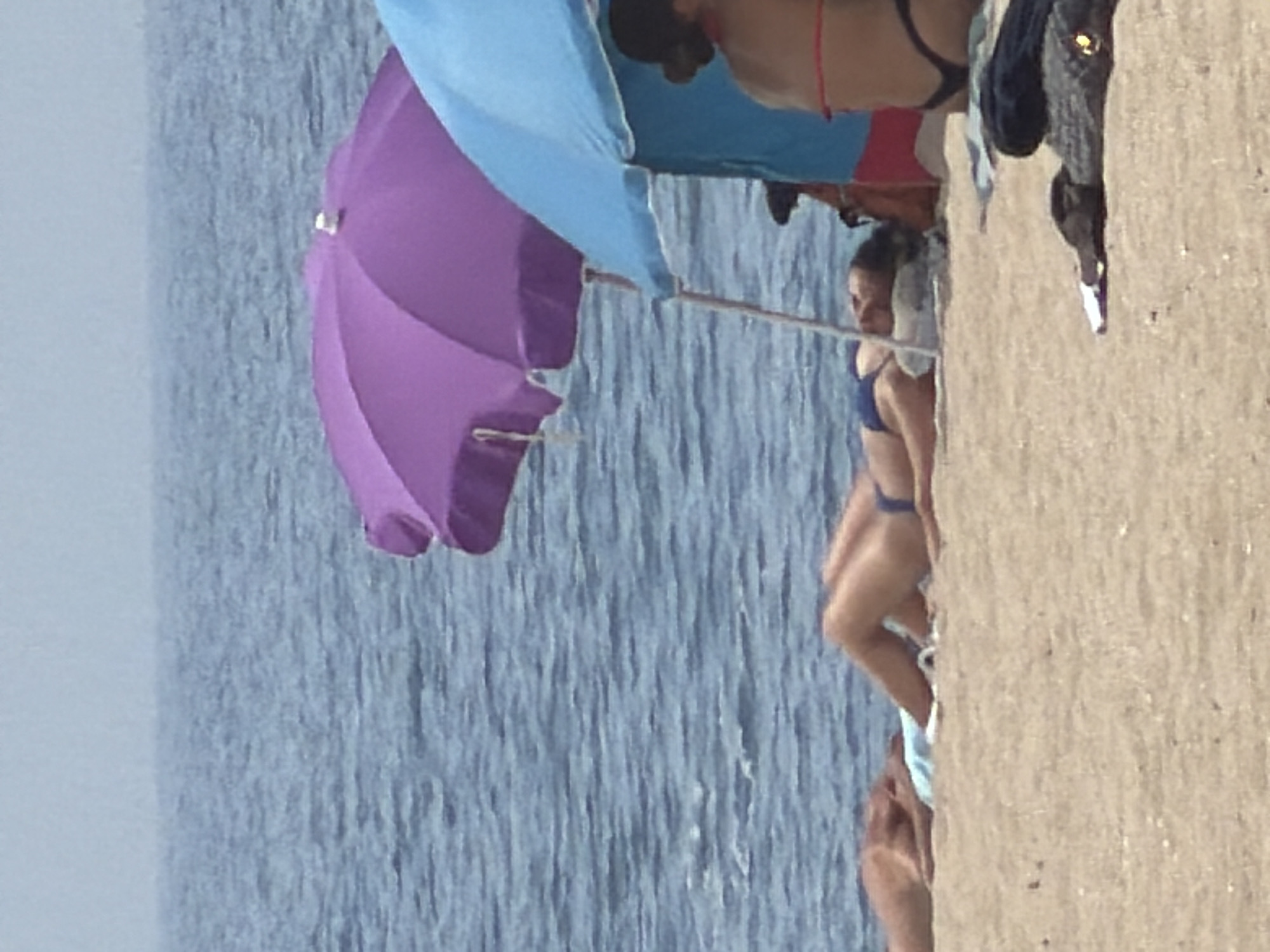 The image depicts a beach scene in Espinho, Portugal. In the foreground, two people are sunbathing on the sand. One individual, lying on their back, is partially obscured by a purple umbrella. Another person is visible further to the right, wearing a bikini, with their back to the camera. A blue umbrella is also present, partially shading the area. The sea is visible in the background, with a hazy sky above. The sand appears light brown with scattered debris. A dark object resembling a folded towel or blanket is in the lower right corner of the image.