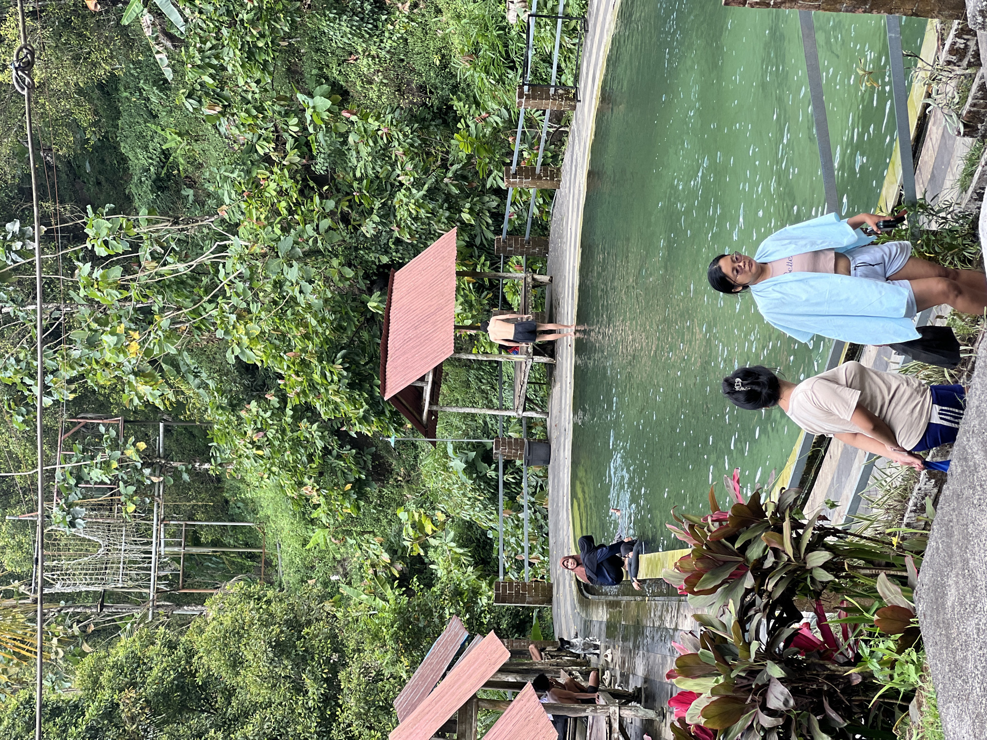 In Sukasada, Bali, Indonesia, a large, emerald-green natural pool is the central focus of this image, nestled within a lush, tropical environment. The pool is surrounded by a paved walkway and a metal railing. Several small, open-air gazebos with red corrugated metal roofs are positioned around the pool's edge, providing shade for visitors.
Several individuals are present. In the middle of the pool, a person with dark hair and wearing black swimwear is partially submerged, with their head and shoulders visible. Near the edge of the pool, a man with dark hair and wearing black shorts stands facing away from the camera, with one foot in the water.
On the paved area to the left, two women stand facing the pool. The woman closer to the foreground has dark hair pulled back in a bun and is wearing a beige t-shirt and dark shorts. The woman in front of her is wearing a light blue cover-up over a pink cropped top and grey shorts, with sunglasses on her head.
Further to the left, under one of the gazebos, several people are seated on a wooden bench. A person with dark hair wearing a headscarf and dark clothing is also seated. A dog with dark fur is visible near the seated individuals.
The background is dominated by dense, green vegetation, with trees and various plants creating a natural, jungle-like atmosphere. In the upper left of the image, a rustic rope structure, possibly part of an adventure course, is visible amidst the foliage. The overall scene suggests a tranquil natural swimming area accessible to the public.