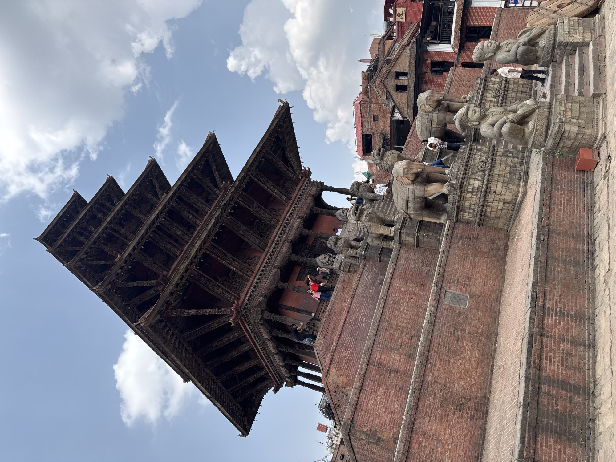 This image depicts the Nyatapola Temple in Bhaktapur, Nepal. It's a five-storied pagoda-style temple with a red brick base and dark wooden support beams. The sky is partly cloudy. People are on the temple's platform and stairs, some standing and some walking. Carved stone statues of animals guard the stairs leading up to the temple. Other buildings can be seen in the background, suggesting an urban setting. The photo appears to have been taken from a slightly low angle, emphasizing the temple's height.