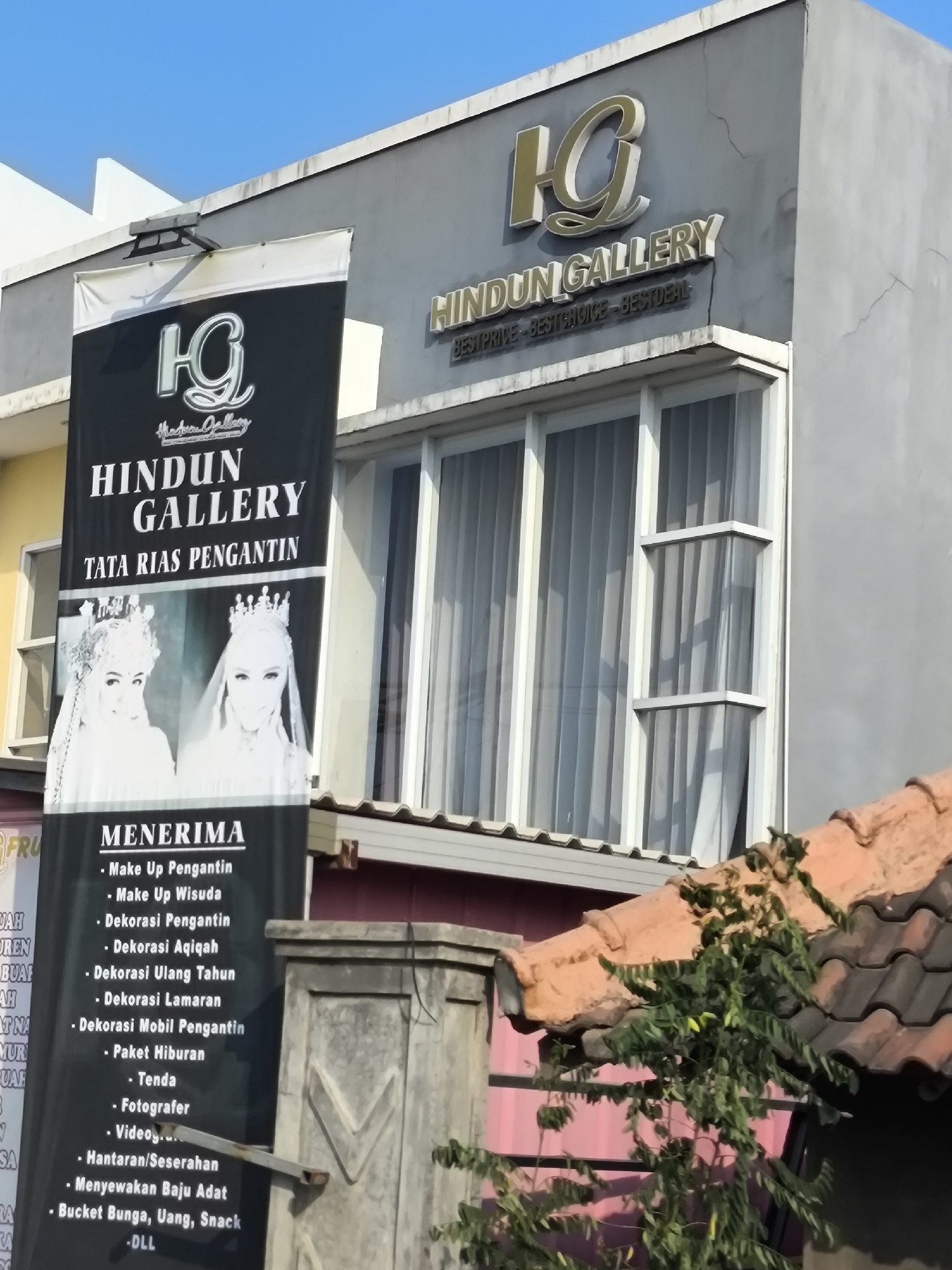 The image shows the exterior of a business called "HINDUN GALLERY" located in Cilegon, Indonesia. A large black banner hangs in front of the building, advertising "TATA RIAS PENGANTIN" (wedding makeup) and listing various services such as "Make Up Pengantin," "Make Up Wisuda," "Dekorasi Pengantin," and "Sewa Baju Adat" (renting traditional attire). The banner features images of two brides in traditional wedding attire. Above the banner, the "HINDUN GALLERY" logo is visible on the building's facade, along with the tagline "BEST PRICE - BEST CHOICE - BEST DEAL." The building has large windows with white curtains. The foreground shows a tiled roof and some greenery. The overall scene suggests a commercial establishment offering wedding-related services.