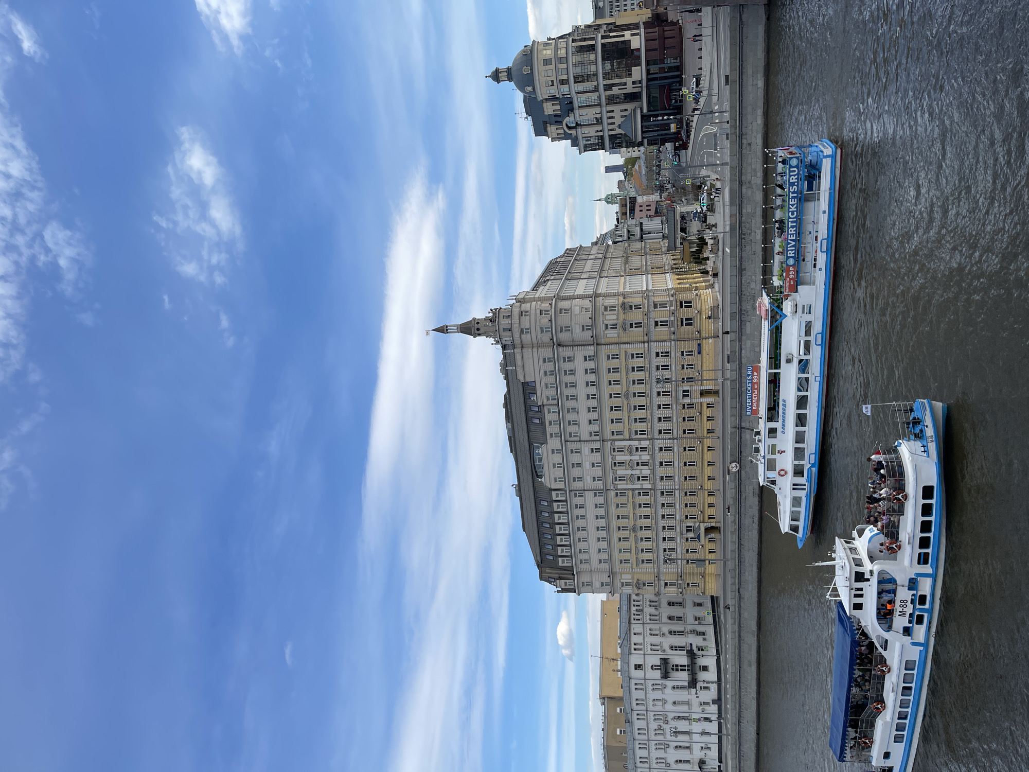 The photograph shows an oblique high-angle view of a city, identified as Moscow, Russia. The sky is blue with streaky white clouds. Notable architectural features include multiple tall buildings. The river below showcases three white riverboats, two of which are filled with people. A building, labeled "RIVERTICKETS.RU," is positioned along the riverbank.