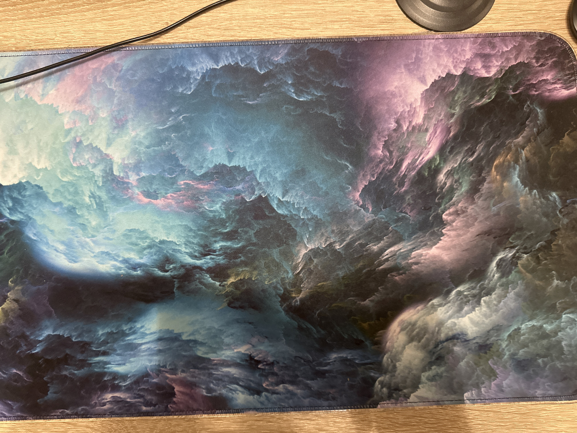 A rectangular fabric mat featuring an abstract, multicolored cloud or nebula design is centrally positioned on a light-colored wooden surface. The mat's pattern consists of intricate swirls and gradients in shades of dark blue, teal, light blue, purple, pink, and subtle accents of yellow and green. Its perimeter is finished with a dark, stitched seam. The underlying wooden surface displays a visible grain, oriented primarily vertically on the left side and horizontally on the right side. A thin, black electrical cable runs diagonally across the upper right portion of the image, partially obscuring the mat's right edge. In the bottom right corner, a segment of a dark gray, circular object with a matte texture rests on the wooden surface adjacent to the mat. No individuals, specific actions, or discernible location within the city of Unknown, Unknown are present.