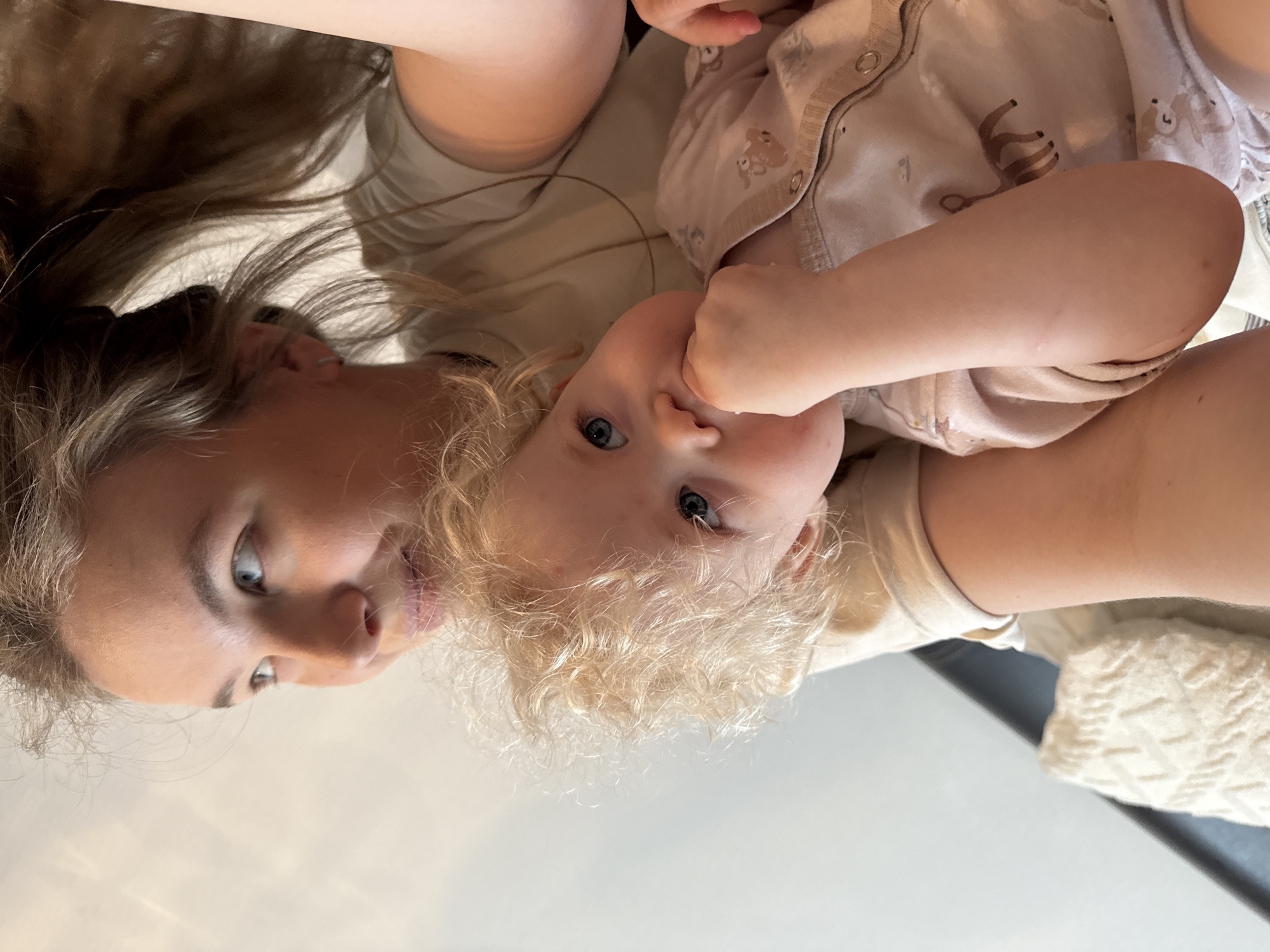 This intimate, close-up portrait captures a woman and a young child, likely a mother and her baby or toddler, in an indoor setting. The scene is located in Vladimir, Russia, based on the provided context. Both subjects are looking directly into the camera, which appears to be positioned above them.
The woman, on the left, has long, light brown hair cascading around her and striking light blue-grey eyes. She wears a plain, light-colored top and exhibits a soft, content expression, conveying affection. The child, on the right, possesses bright blue eyes and a head full of curly blonde hair. Their left hand is brought up towards their mouth, a common gesture for a young child. The child is dressed in a light-toned onesie featuring subtle, faded patterns of what appear to be deer or similar animals.
The pair are positioned closely, suggesting a tender, familial bond, with the child possibly being held or resting on the woman. The background is intentionally blurred, presenting soft, neutral tones that indicate an indoor environment bathed in gentle, natural daylight. A textured, white knitted fabric or blanket can be seen partially on the far right. The overall atmosphere of the image is warm and intimate, highlighting a moment of connection. There is no visible text present in the image.