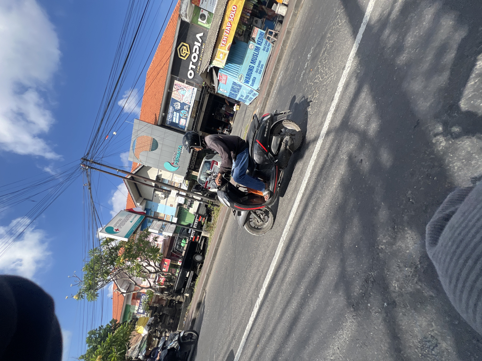 The image captures a street scene in Denpasar, Indonesia, under a bright, partly cloudy sky. A man wearing a black helmet, dark long-sleeved shirt, and blue jeans is riding a black scooter. He is in motion, heading towards the right side of the frame, with his right foot resting on the footrest and his left foot slightly off. The scooter has the word "SCOOPY" visible on its side.
The background features a row of buildings with orange tiled roofs. Several signs are visible, including "APOTEK PERMATA" with a green logo, "OTOPEDIA COATING & DETAILING" with a golden logo, and "TEHTJAP SOLO". Other signs are partially obscured. A black pickup truck with "MITS BISHT" written on its side is parked along the street. Other vehicles and elements of a busy urban environment, such as power lines and trees, are also present. The road is paved and marked with a white lane divider. Shadows of trees and possibly power lines are cast across the asphalt. In the foreground, a blurry gray object, likely part of the photographer's clothing, is visible.