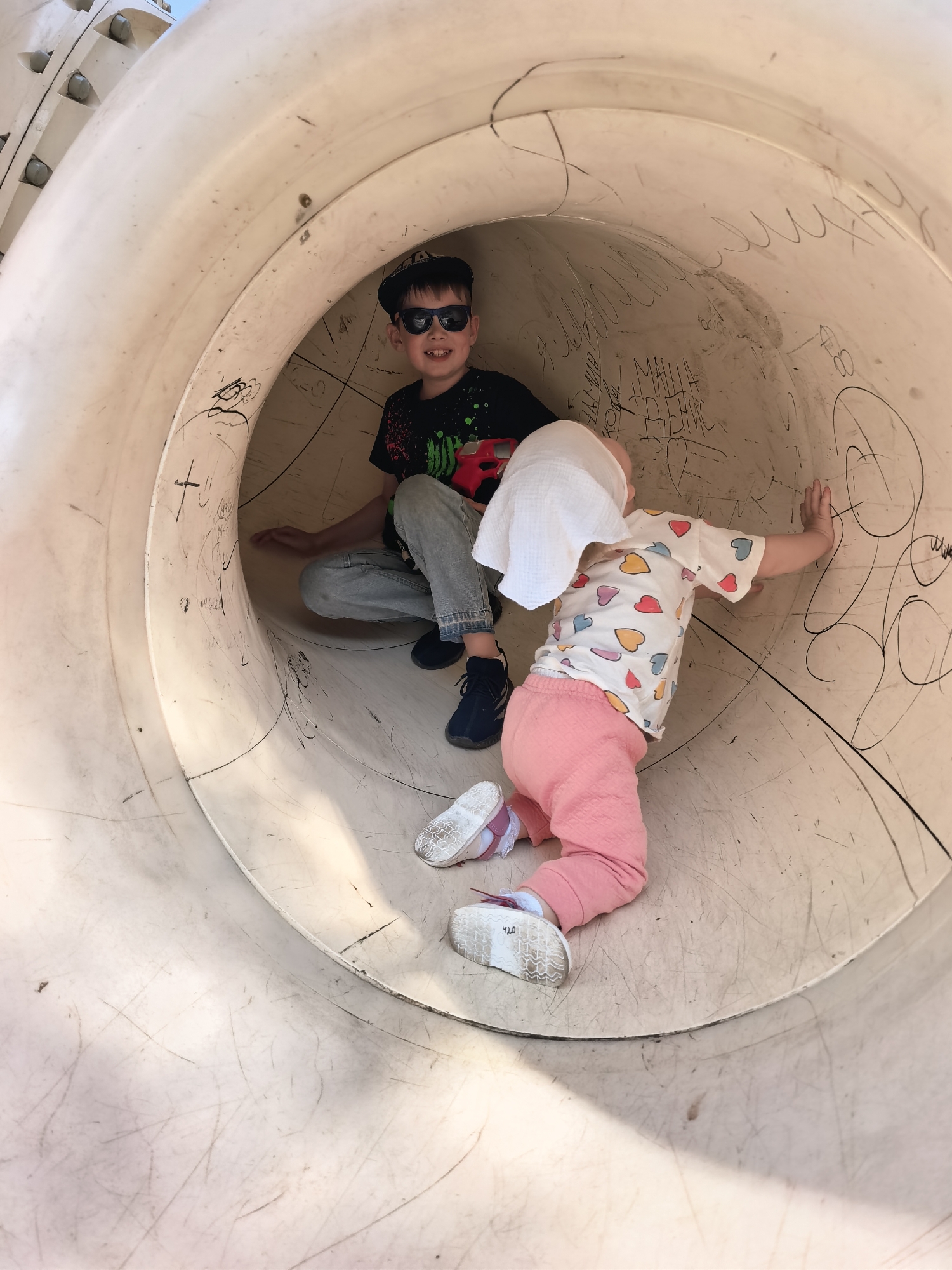 Two children are inside a large, white, cylindrical tunnel, possibly part of a playground slide. The tunnel appears to be made of plastic and is marked with various scribbles and scratches.
In the foreground, a toddler is on their hands and knees, facing away from the camera. They are wearing a white head covering, a white t-shirt with colorful hearts, and pink leggings. Their feet are clad in white sneakers with pink accents.
Further back in the tunnel, a young boy is sitting and looking towards the camera. He is wearing a black baseball cap, sunglasses, and a black t-shirt with a colorful graphic. He is wearing blue jeans and dark sneakers. He is holding a red toy in his left hand.
The lighting suggests an outdoor setting, with sunlight casting shadows within the tunnel. The image was taken in Sterlitamak, Russia.