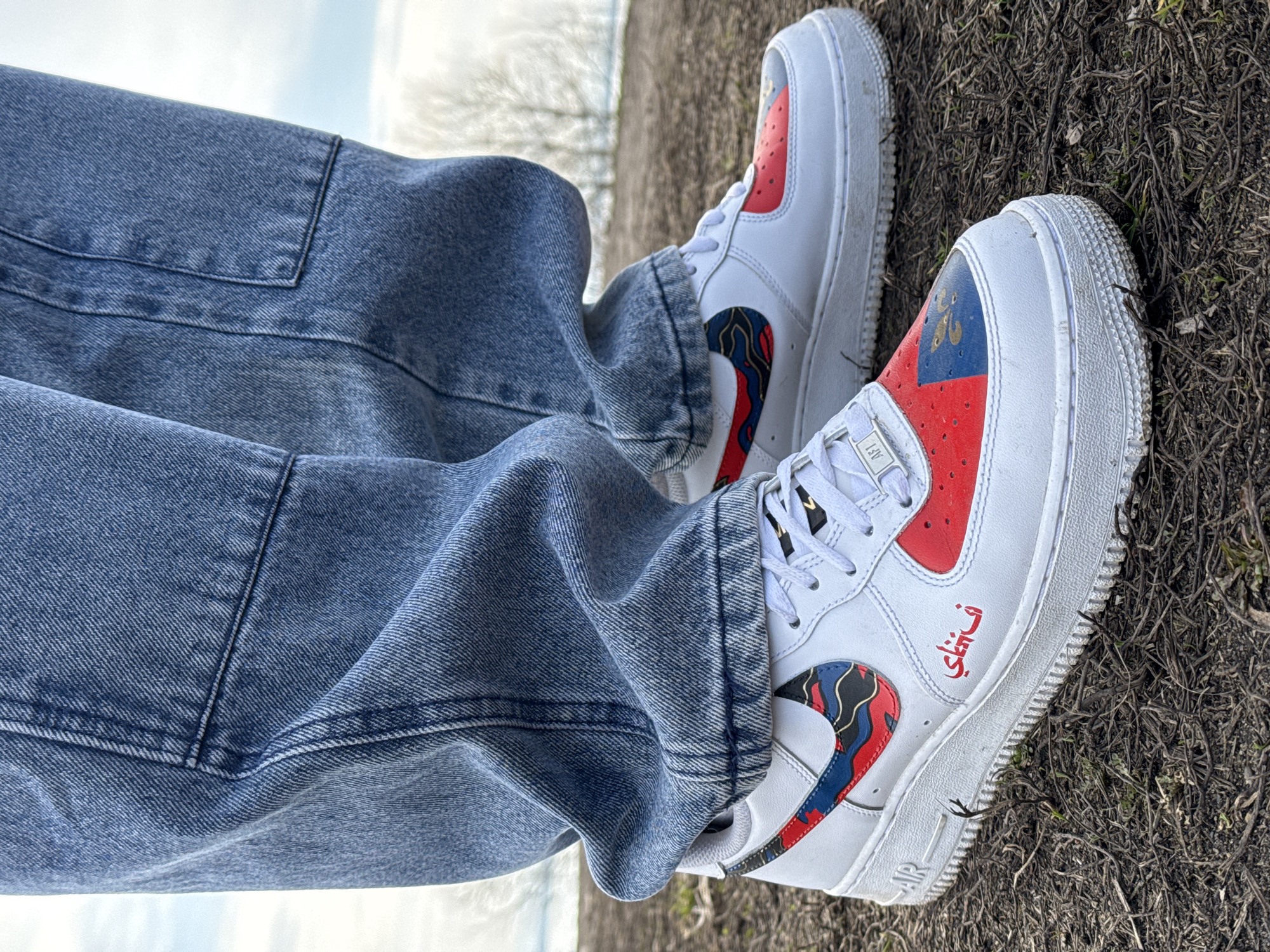 A person wearing baggy, light blue denim pants and custom white Nike Air Force 1 sneakers is seated outdoors on a patch of dirt and dry grass. The sneakers feature red and blue accents with a swirling pattern within the Nike swoosh and red detailing on the toe box. One shoe has red Arabic lettering near the sole. The sky is overcast, and bare trees are visible in the background. The photograph is taken from a low angle, looking up at the subject. Location: Prasolovo, Russia.