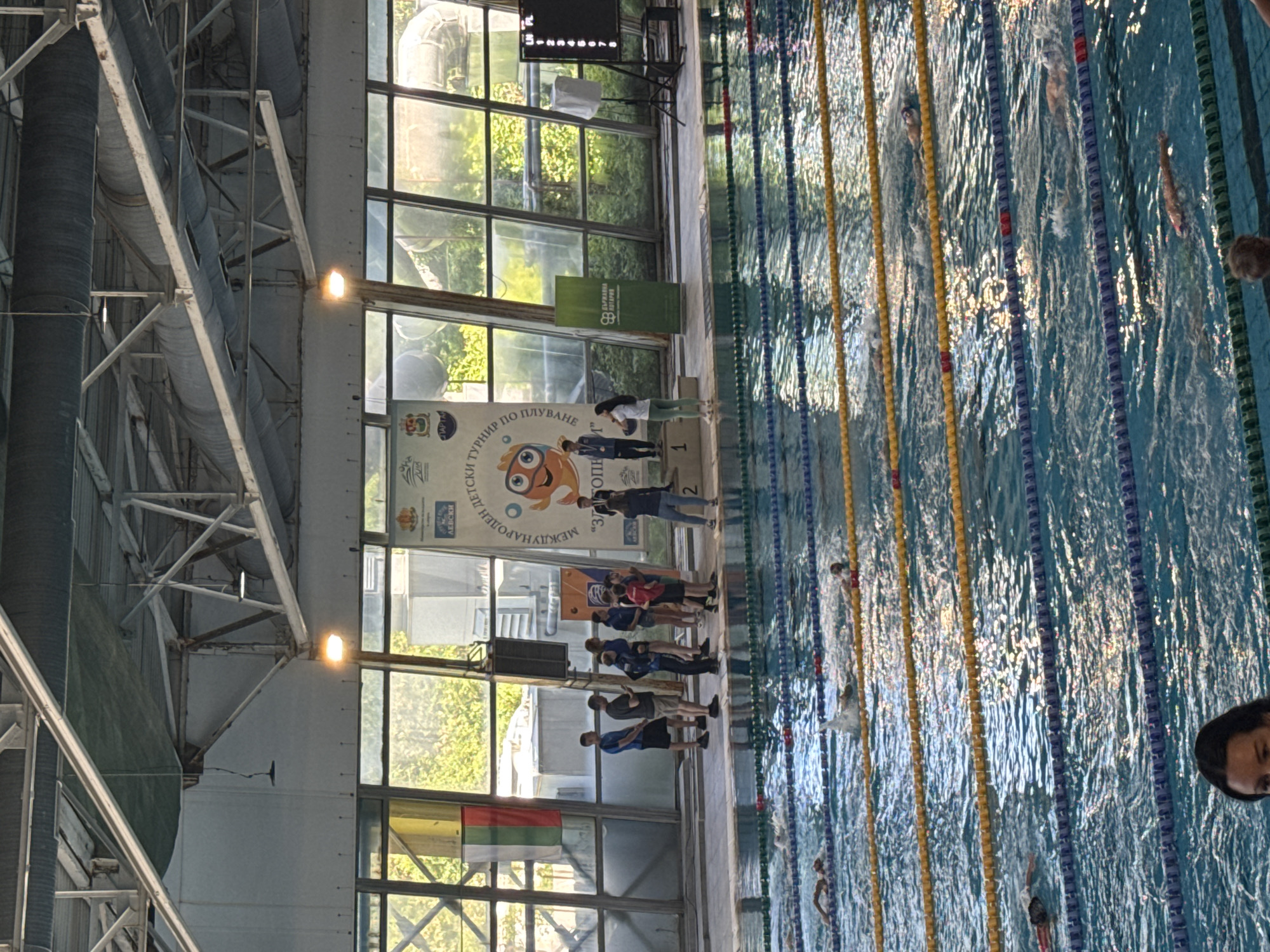 The image shows an indoor swimming pool in Sofia, Bulgaria, possibly during a competition. Swimmers are in the lanes. Several people are standing near the starting blocks. A podium for awards is visible, as well as banners with the name of the competition. The pool is surrounded by large windows. The ceiling is made of metal beams.