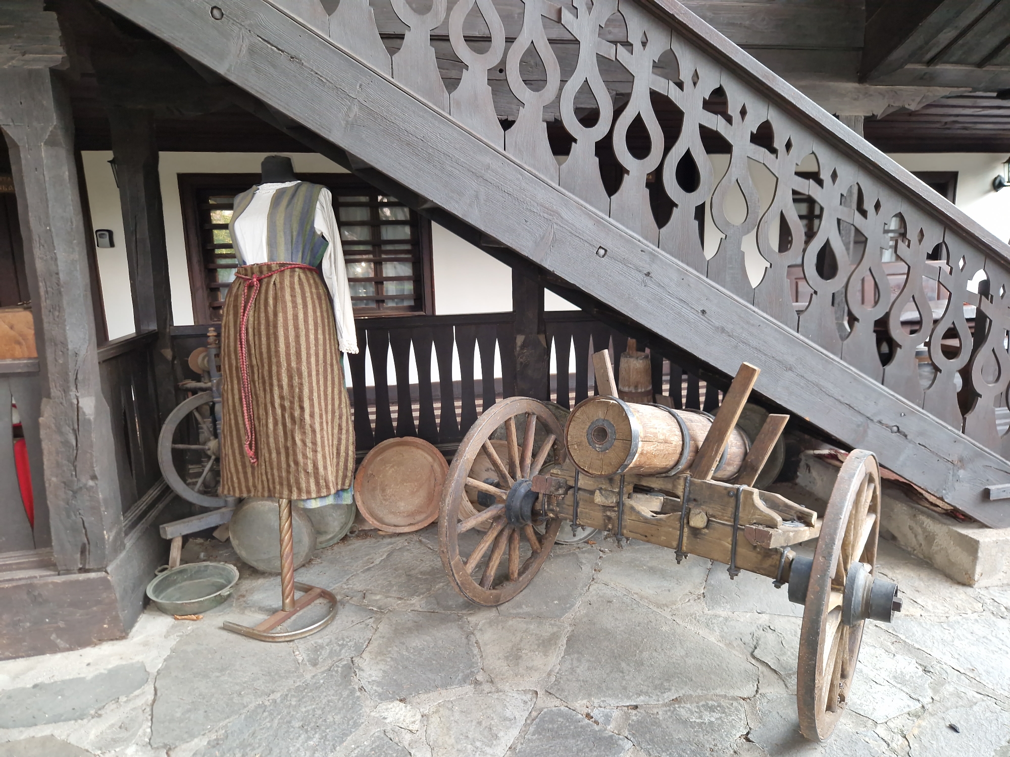 The image shows an outdoor display of traditional Bulgarian folk costumes and historical artifacts, likely at an open-air museum or cultural heritage site in Panagyurishte, Bulgaria. In the foreground, a wooden cart with large spoked wheels and a barrel-like structure is positioned on a stone-paved ground. To the left, a mannequin is dressed in a traditional striped skirt, a white blouse, and a vest, cinched with a red rope belt. Behind the mannequin, a spinning wheel and a metal basin are visible. The background features a wooden veranda with ornate railings and a building with traditional architecture, including wooden beams and windows. The overall scene evokes a sense of historical preservation and cultural display, showcasing elements of Bulgarian rural life and craftsmanship.