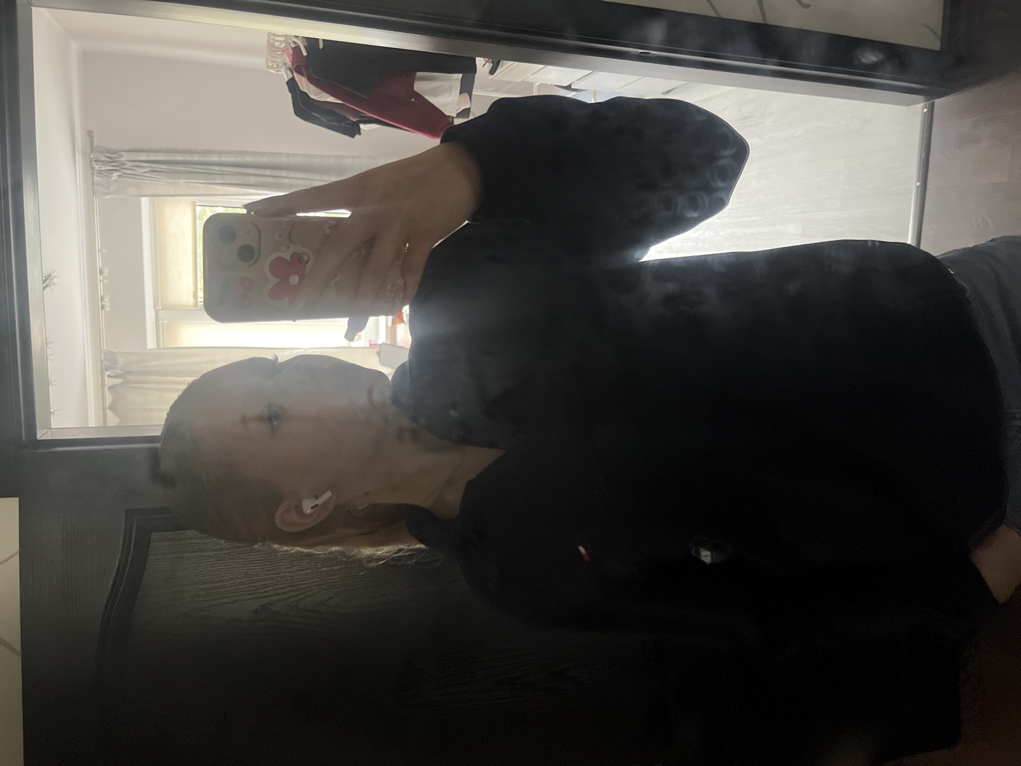 A young woman is reflected in a mirror, taking a selfie with her phone. She has blonde hair pulled back and is wearing a dark navy sweatshirt. She has white earbuds in her ears. The background of the reflection shows a room with light-colored curtains and a window. To the right, a rack of clothes is visible. The location is a room in a residential apartment in the city of Perm, Russia.