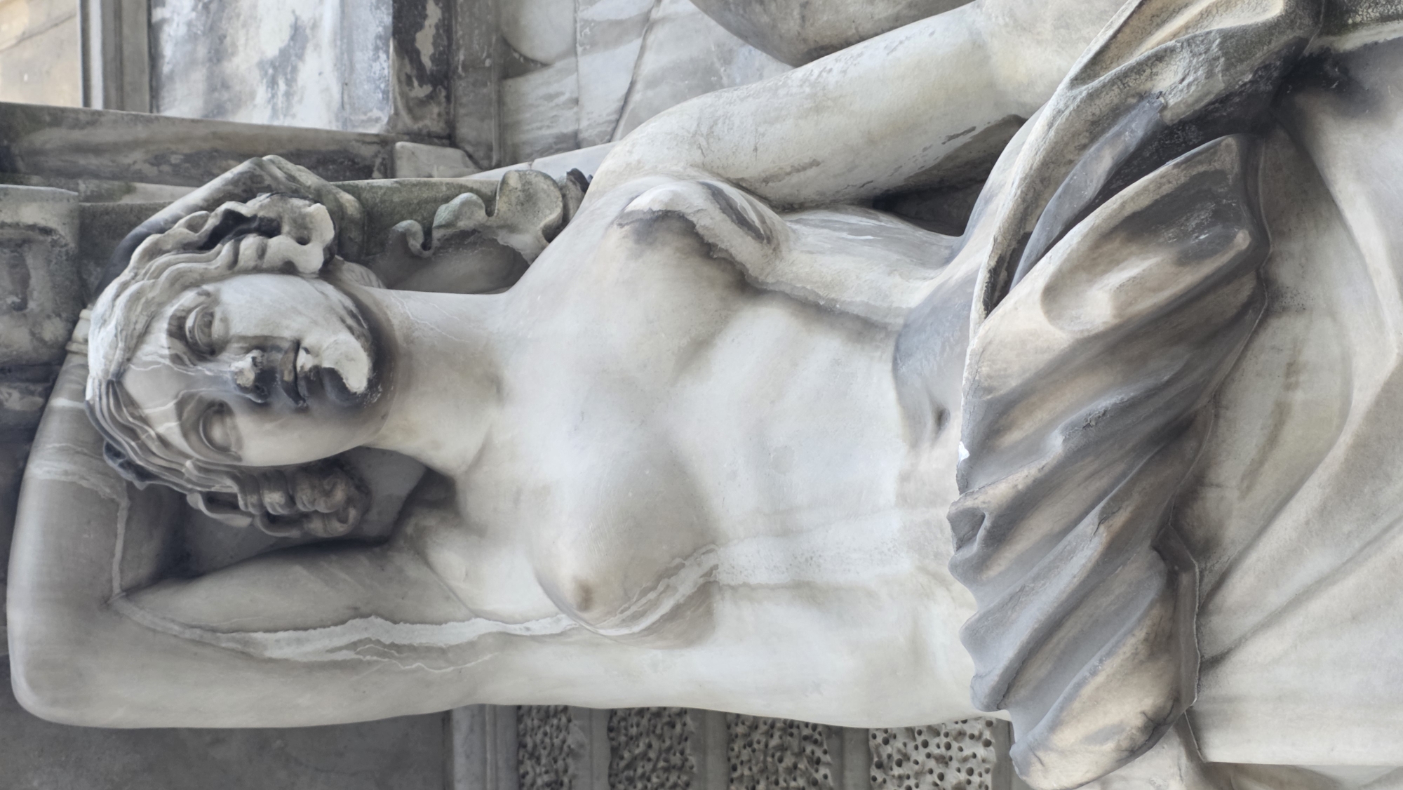 The image depicts a close-up view of a reclining marble statue of a nude female figure. The statue is likely outdoors, showing signs of weathering and discoloration. The statue is located in Saint Petersburg, Russia.