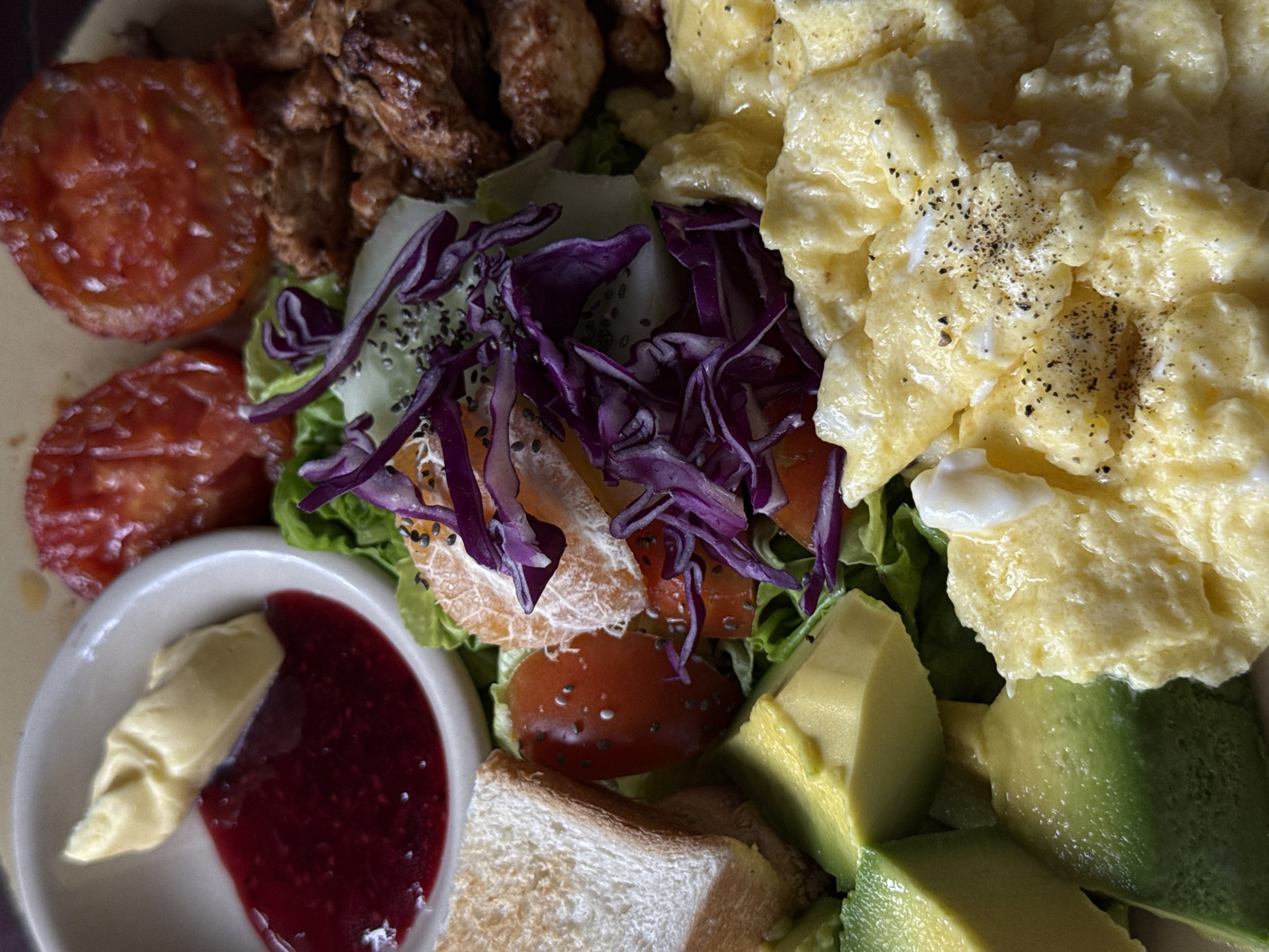 A close-up, overhead shot reveals a plate of food, potentially breakfast, in North Kuta, Indonesia. The plate features scrambled eggs seasoned with pepper, sliced avocado, grilled tomatoes, pieces of fried chicken or similar protein, mixed greens with red cabbage and orange segments, a dish of butter and jam, and a slice of toast. The ingredients are densely arranged, suggesting a hearty meal.