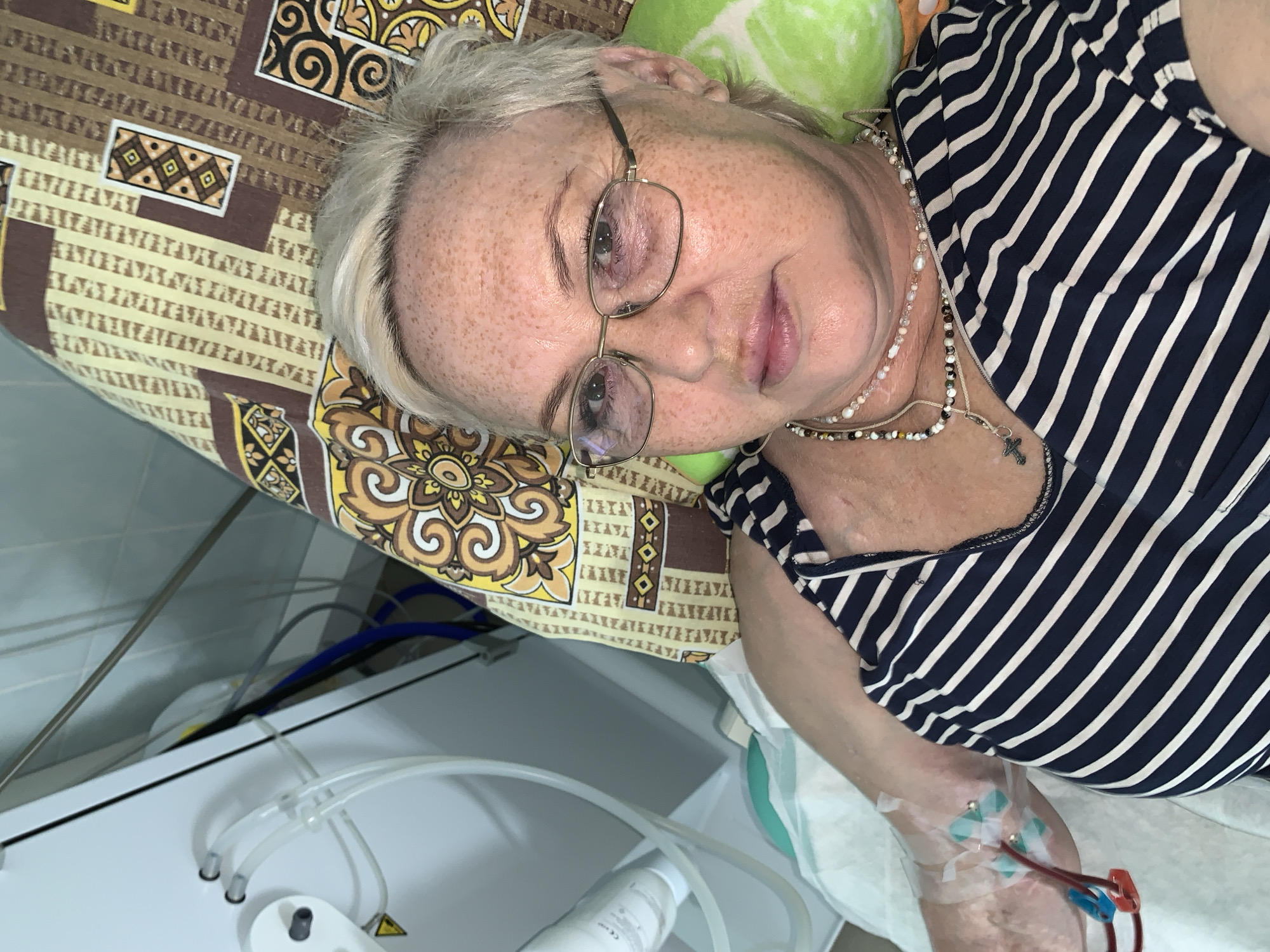 A middle-aged woman with grey hair and freckled skin is lying on a bed or examination table. She is wearing glasses, a striped shirt, and several necklaces, one of which has a cross pendant. There is a pillow or cushion under her head. An intravenous line is visible in her arm, with tubing and medical equipment nearby. The bed has a patterned cover with brown, yellow, and beige designs. The location appears to be a medical facility, based on the equipment present. The location of the image is Chaikovsky, Russia.