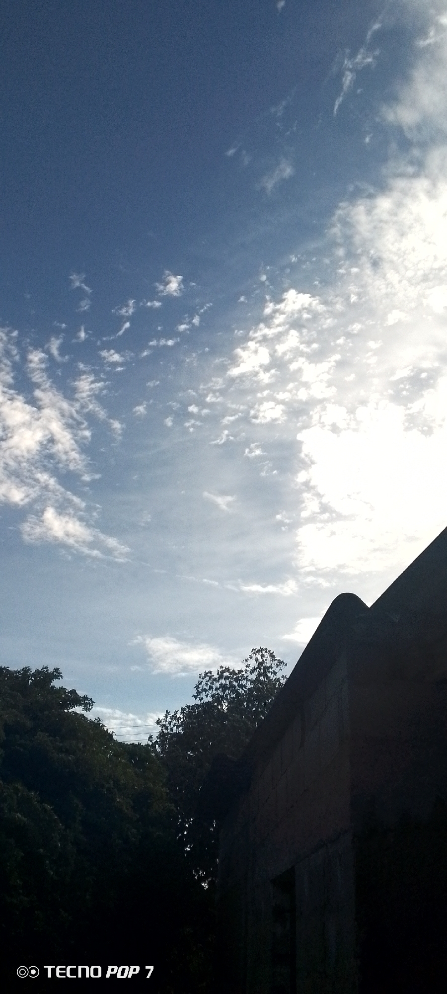 The image depicts a daytime scene in Altagracia, Venezuela, showcasing a view of the sky with scattered white clouds against a blue backdrop. The sun appears to be shining brightly, creating a diffused glow in the upper right portion of the frame. A section of a building or structure, possibly made of concrete blocks, is visible in the lower right corner, partly obscuring the view. Dense foliage from trees is visible on the left side of the frame. "TECNOS POP 7" appears in the lower-left corner, indicating the device used to capture the image.