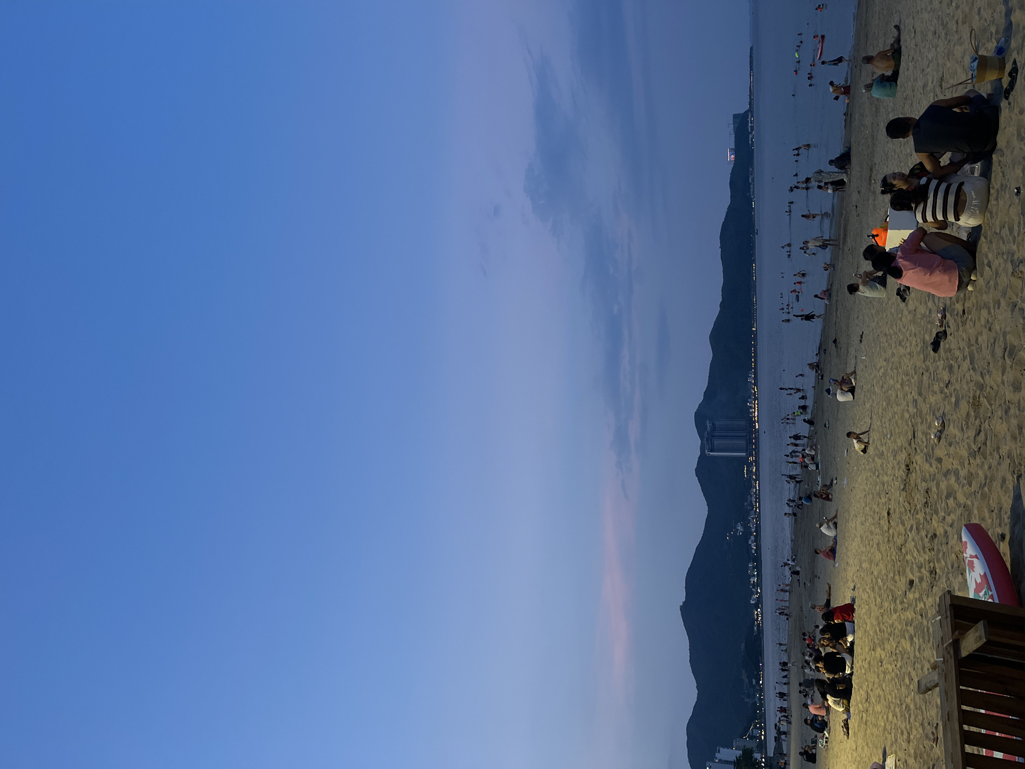 This image depicts a twilight scene on a sandy beach. A vast expanse of clear blue sky dominates the upper portion of the frame, gradually transitioning to a softer blue with hints of pink and purple near the horizon. A range of dark, silhouetted mountains forms the backdrop, with city lights twinkling along the coastline and tall buildings visible on the slopes. The sea is calm and appears populated with numerous people swimming and wading. The foreground is a sandy beach where many individuals are seated or walking. Several groups of people are gathered in conversation, some appearing to be families or friends. Objects on the beach include a wooden structure in the lower left, a red and white kayak, and a woven basket. The overall atmosphere is one of a relaxed evening gathering by the sea.