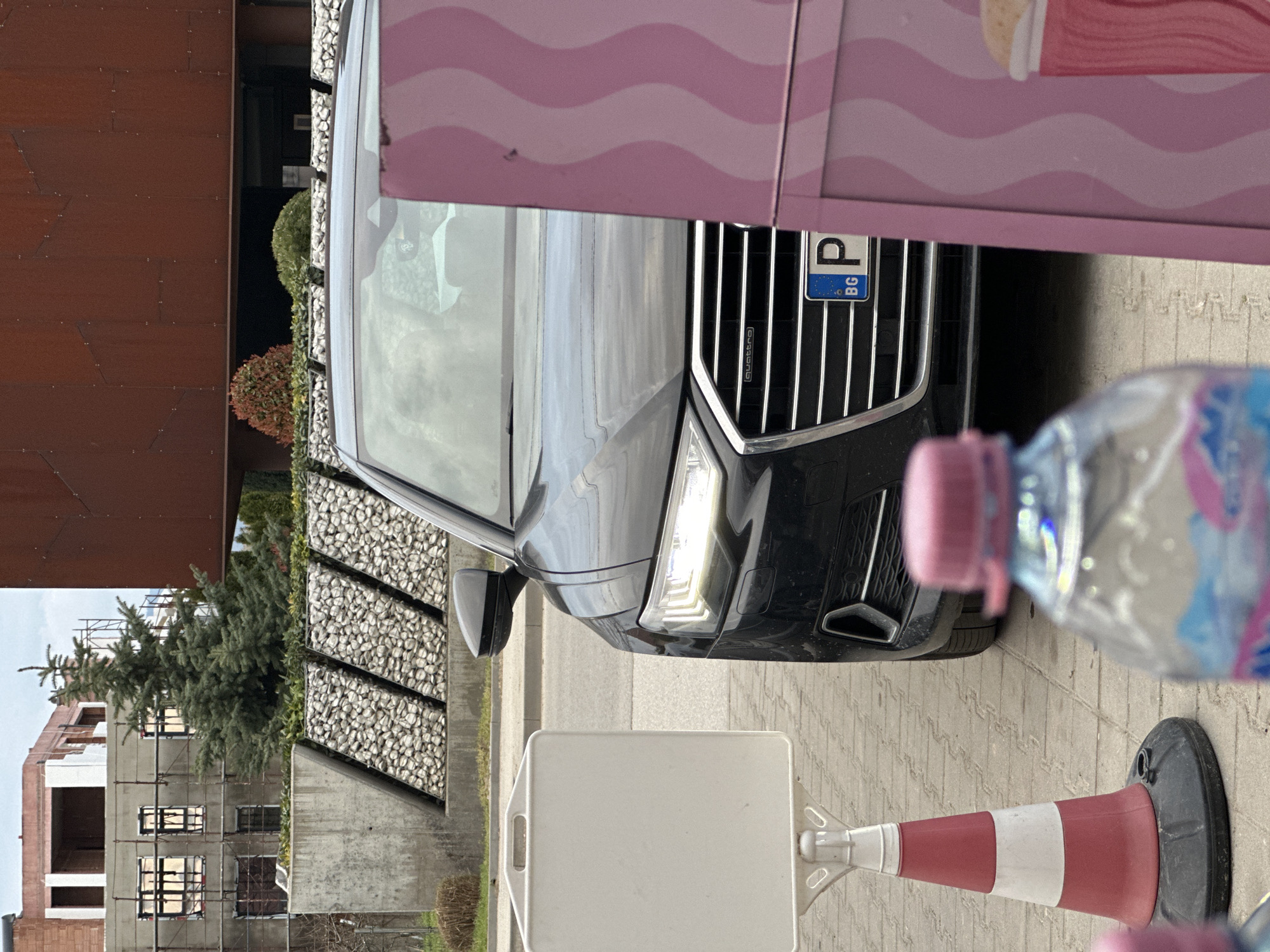 A medium shot depicts a black Audi SUV partially obscured by a pink and white structure, and a plastic water bottle and traffic cone. The car displays a Bulgarian license plate "P" over "BG," indicating it is registered in Plovdiv, Bulgaria. The background reveals a modern building under construction with scaffolding, a concrete and rock wall, and landscaping. The setting is an urban environment on a paved surface.
